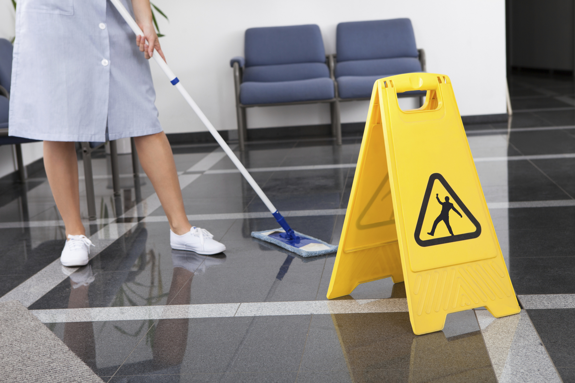 Cleaner mopping a wet lobby floor beside a yellow caution sign and chairs