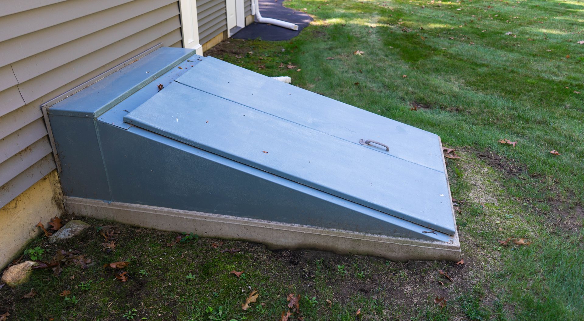 A grey metal cellar door slanted against the beige siding of a house on a grassy lawn.