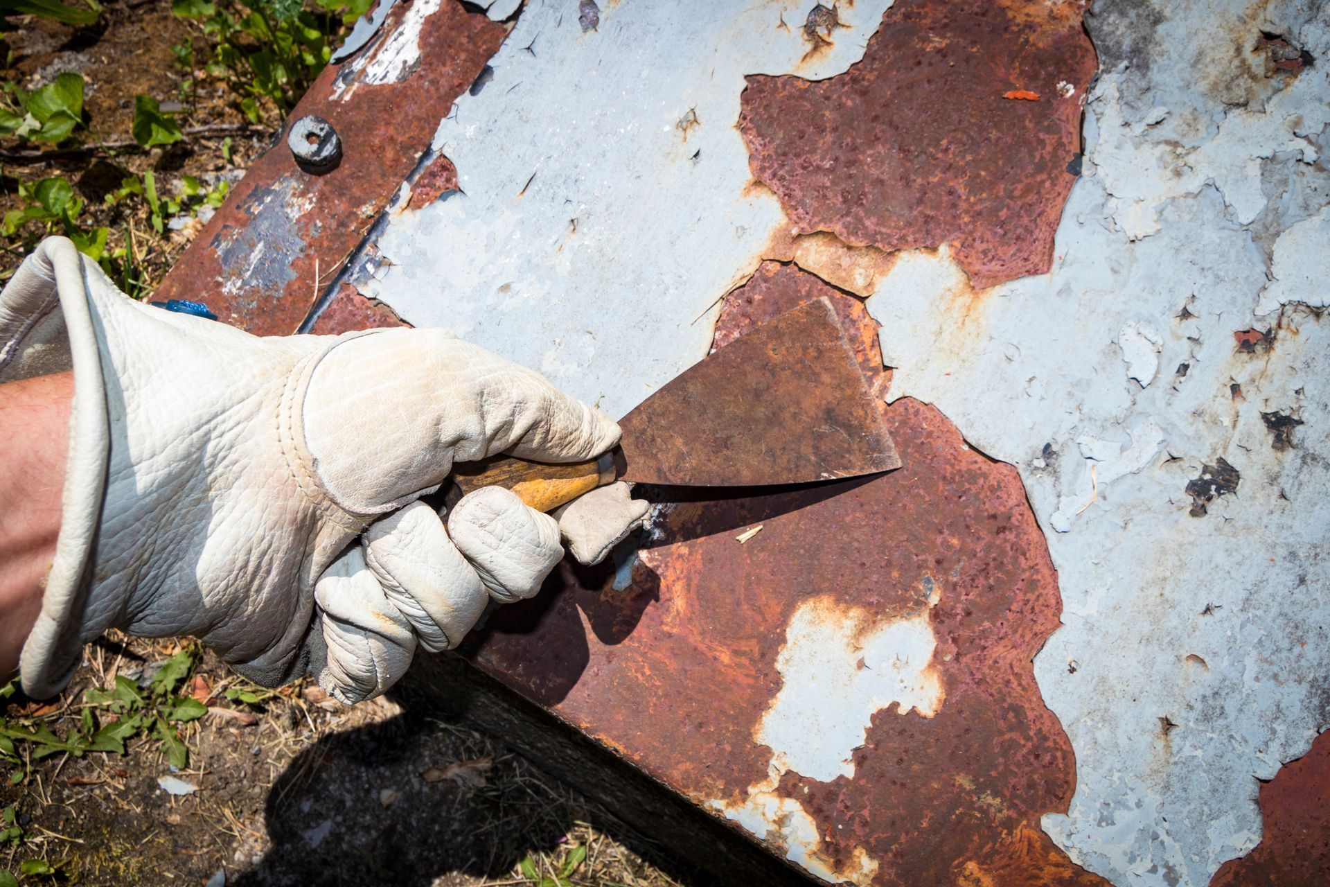 A gloved hand uses a putty knife to scrape peeling paint and rust from a weathered metal surface.