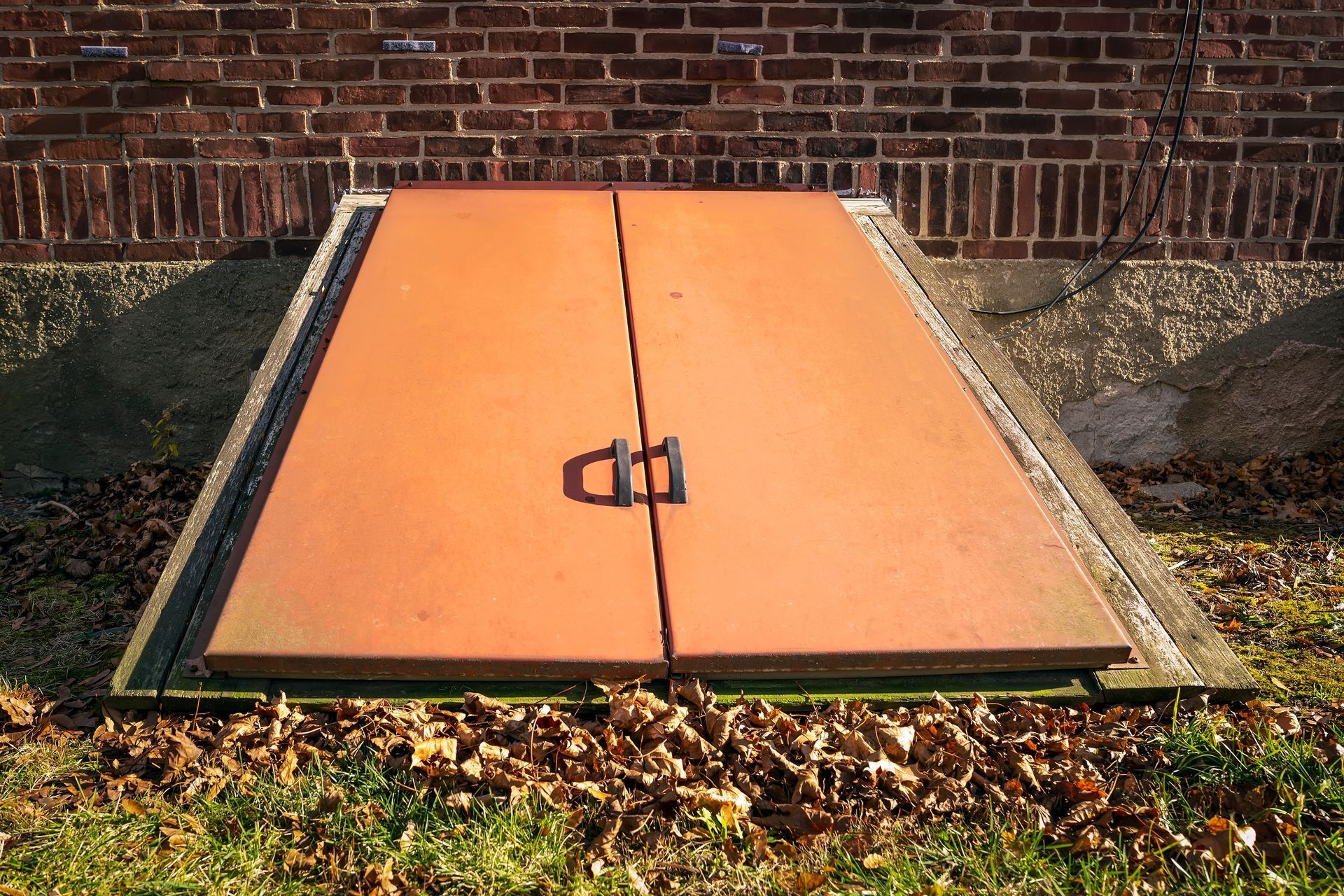 An orange, two-door basement storm cellar entrance set into the ground against a red brick house foundation.