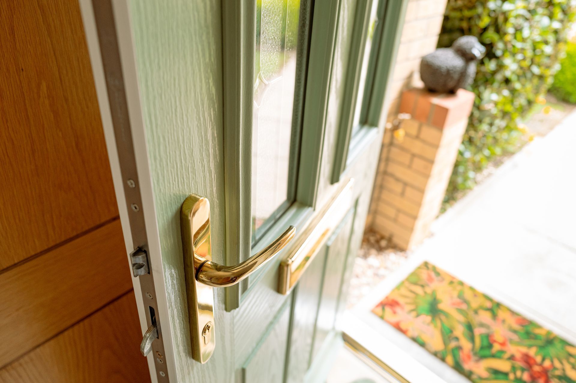 A sage green front door standing slightly ajar, showing a gold-colored handle and an entryway with a rug and brick pillar.