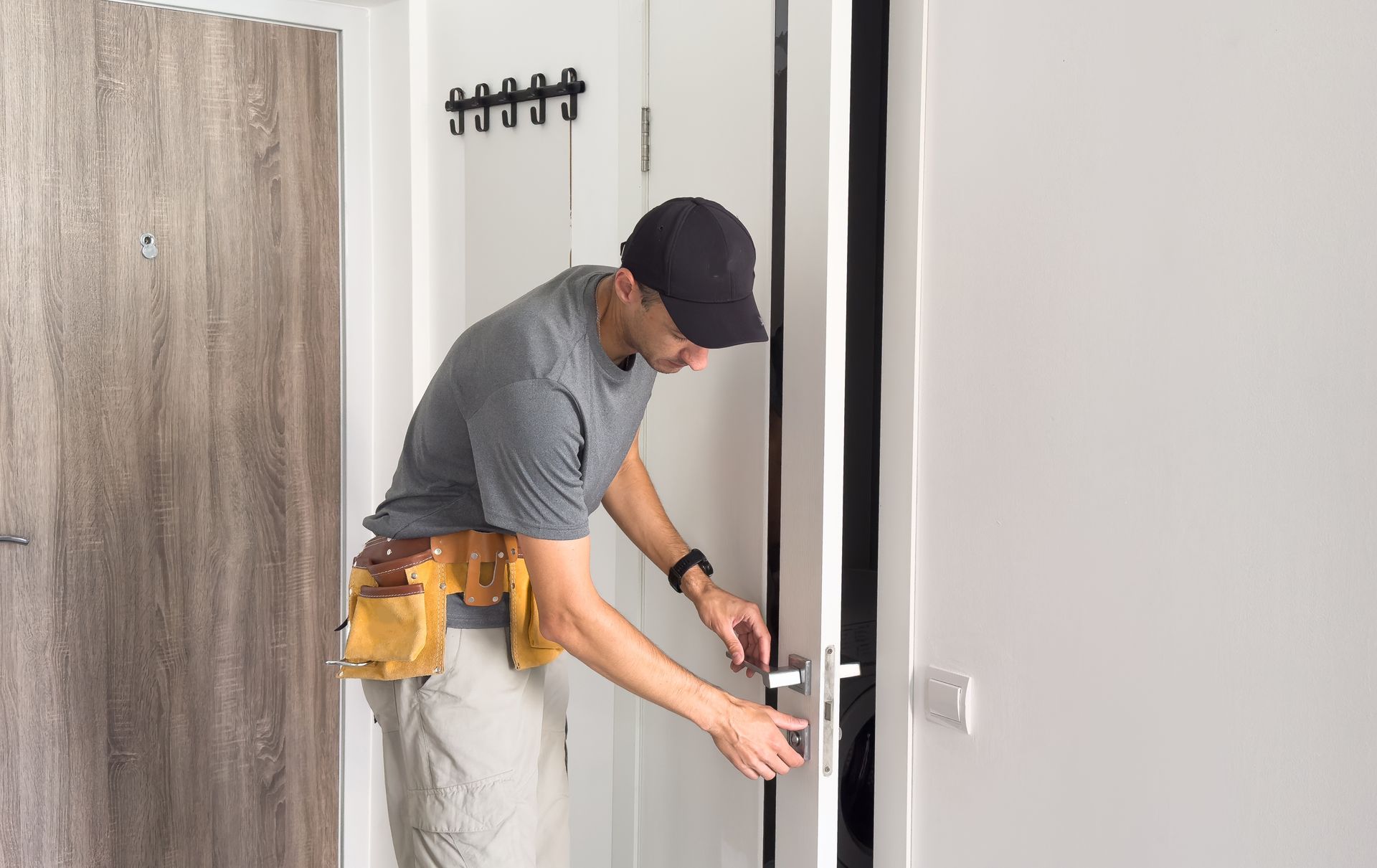 A person in a gray shirt and tool belt installing a handle on a white door in a bright, modern interior.