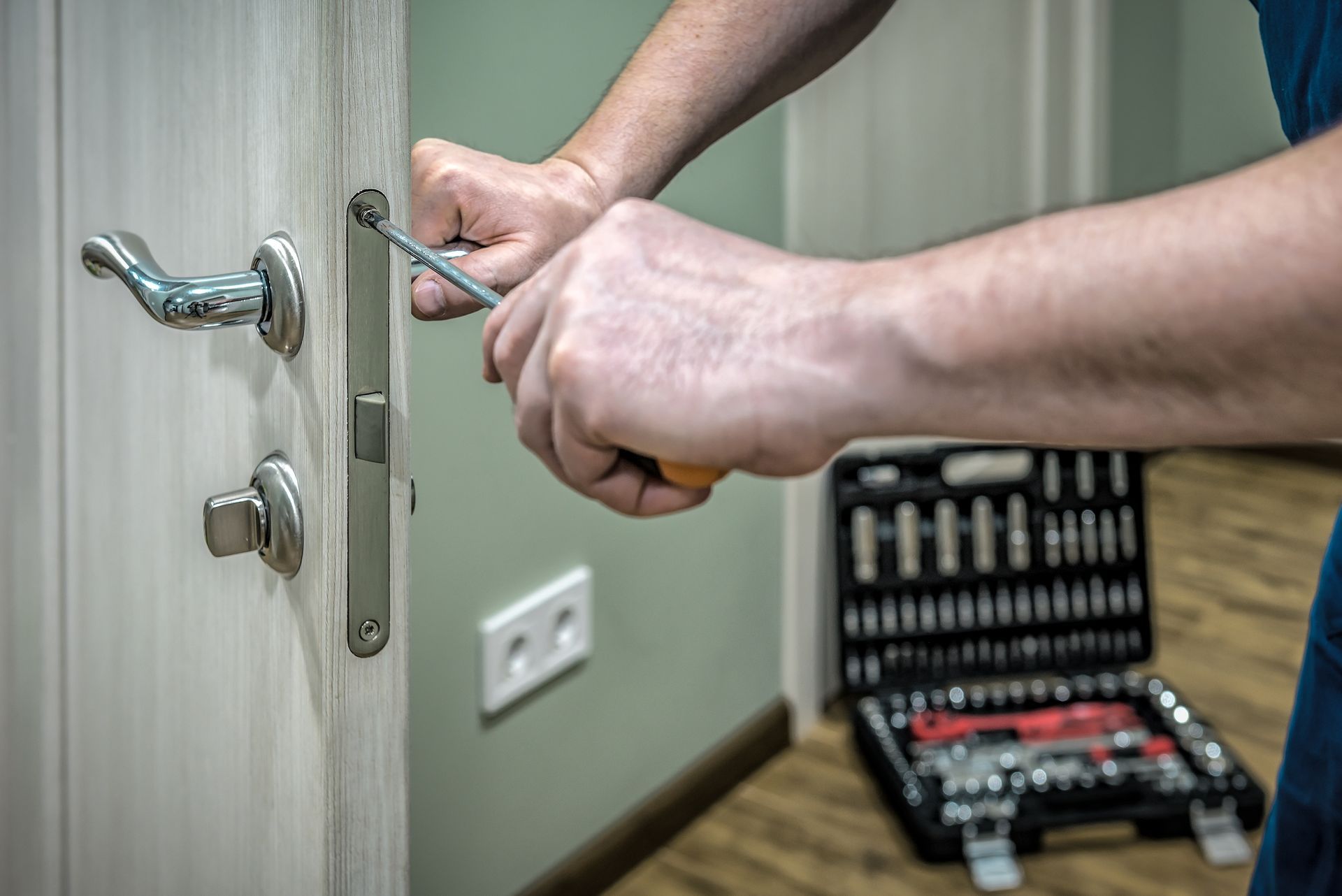 A close-up view of a person using a screwdriver to install or repair a metallic door lock mechanism on a light wood door.