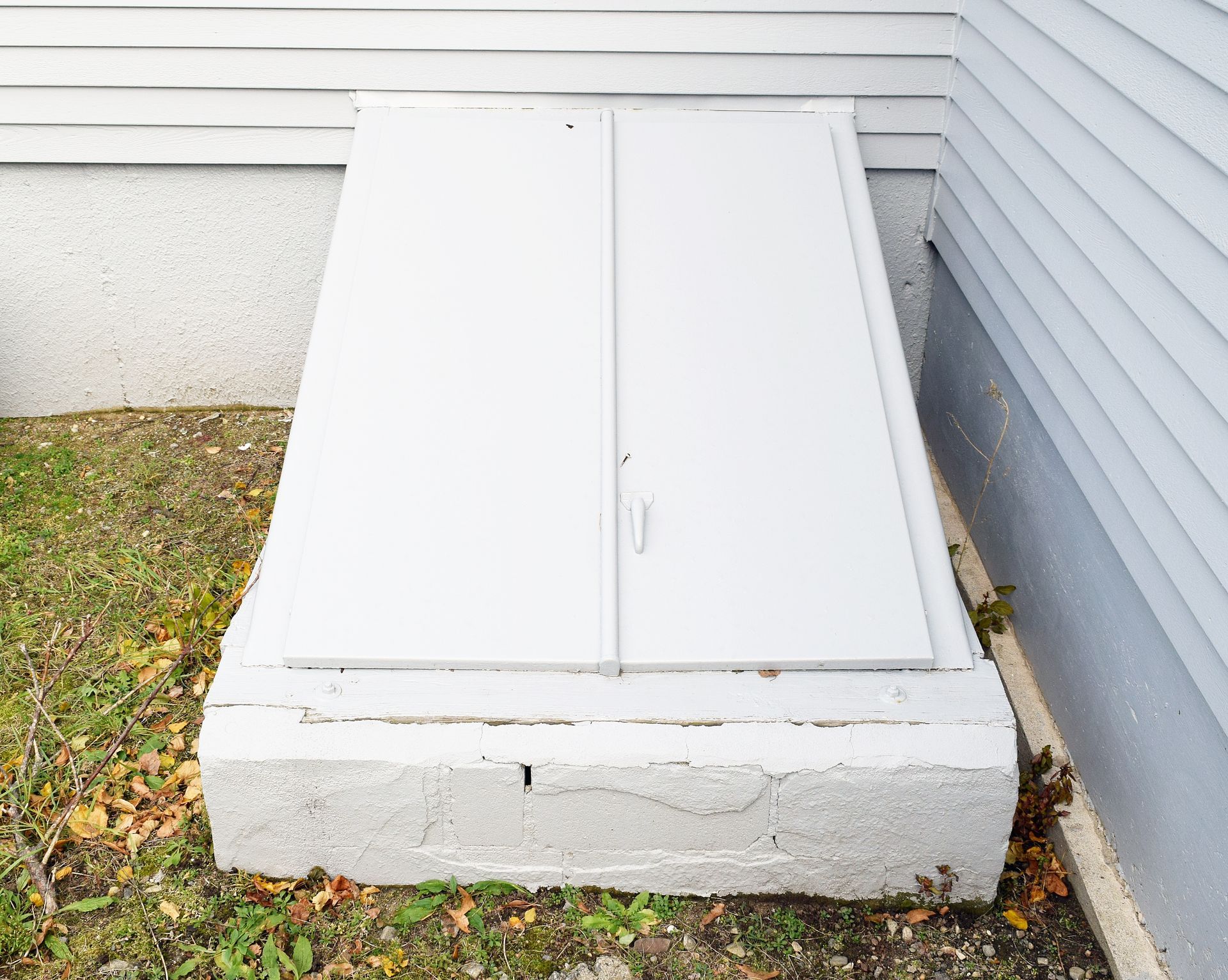 A white metal cellar door angled against the side of a house with grey siding and a concrete foundation.