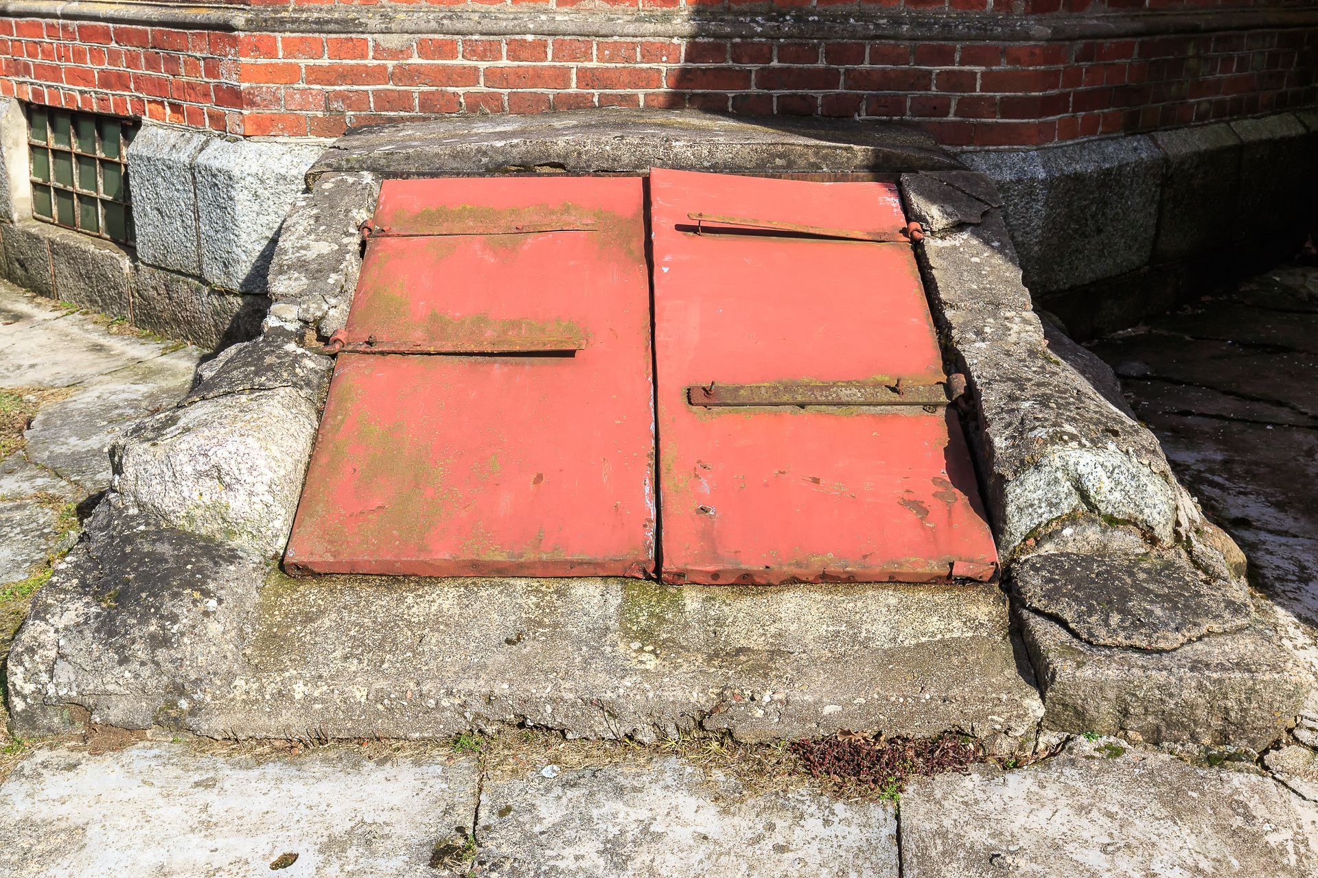 Red, double-door cellar entrance set in a weathered concrete frame against a brick building foundation.