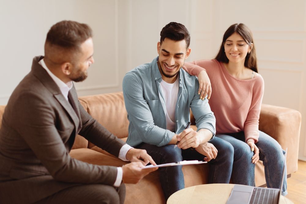 A man in a suit shows a document to a smiling couple on a couch, indoors.