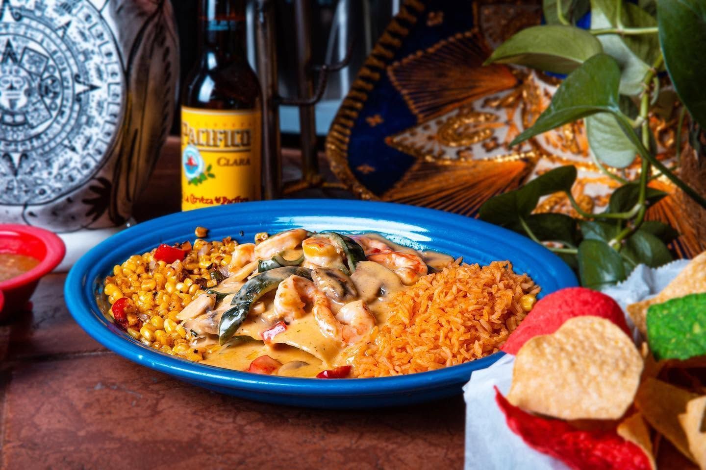 Blue-rimmed plate of saucy enchiladas with rice, beans, and garnish on a table with drinks and bread