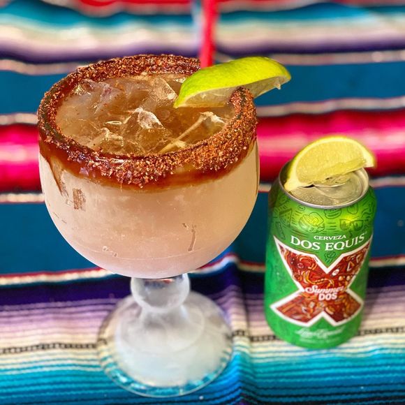 Coconut cocktail with chili-salt rim and lime wedge beside a Dos Equis can on a colorful striped cloth