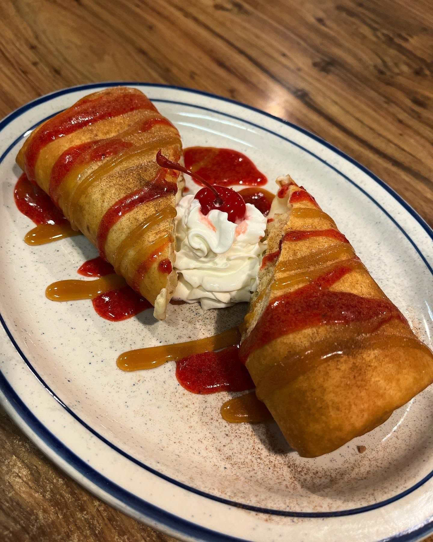 Two rolled fried pastries drizzled with red and caramel sauce, filled with whipped cream on a white plate.