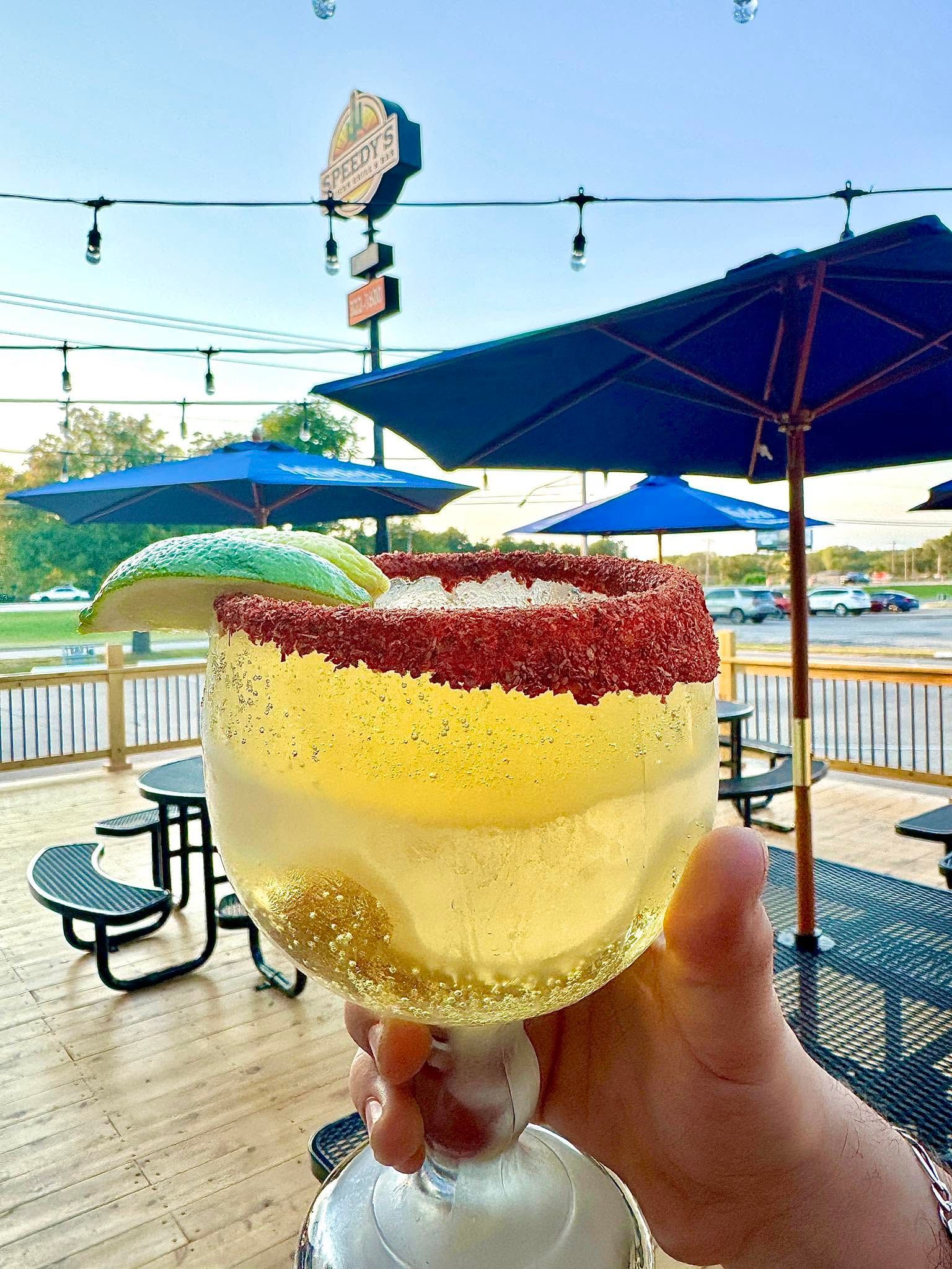 Hand holding a yellow cocktail with a red sugared rim on a sunny waterfront deck with blue umbrellas
