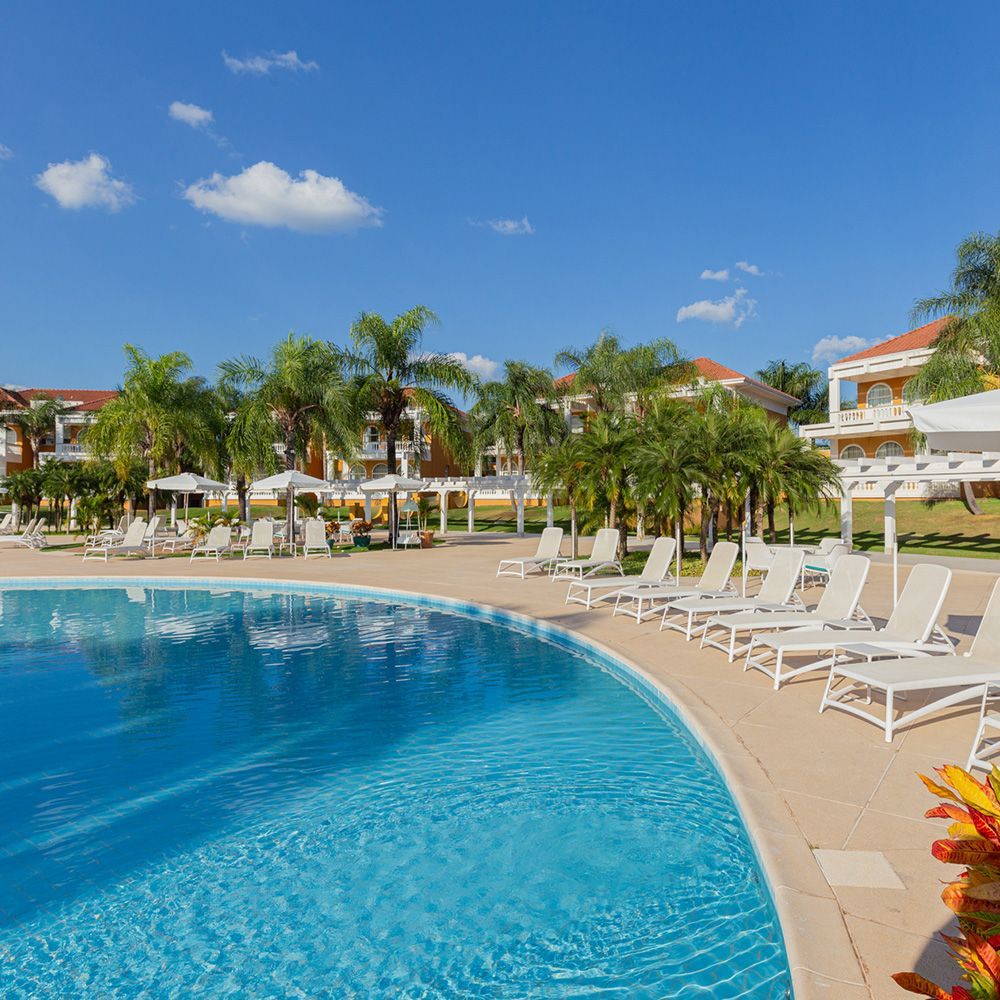 Piscina com espreguiçadeiras e palmeiras em um resort sob um céu azul.