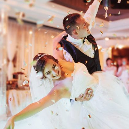 Noivos dançando, confete caindo. Ela está inclinada para trás, sorrindo, e ele a segura no colo. Recepção de casamento.