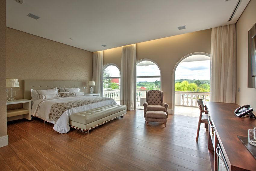 Quarto com cama, janelas em arco, poltrona, escrivaninha e piso de madeira. Paredes bege, luz natural e vista para a varanda.