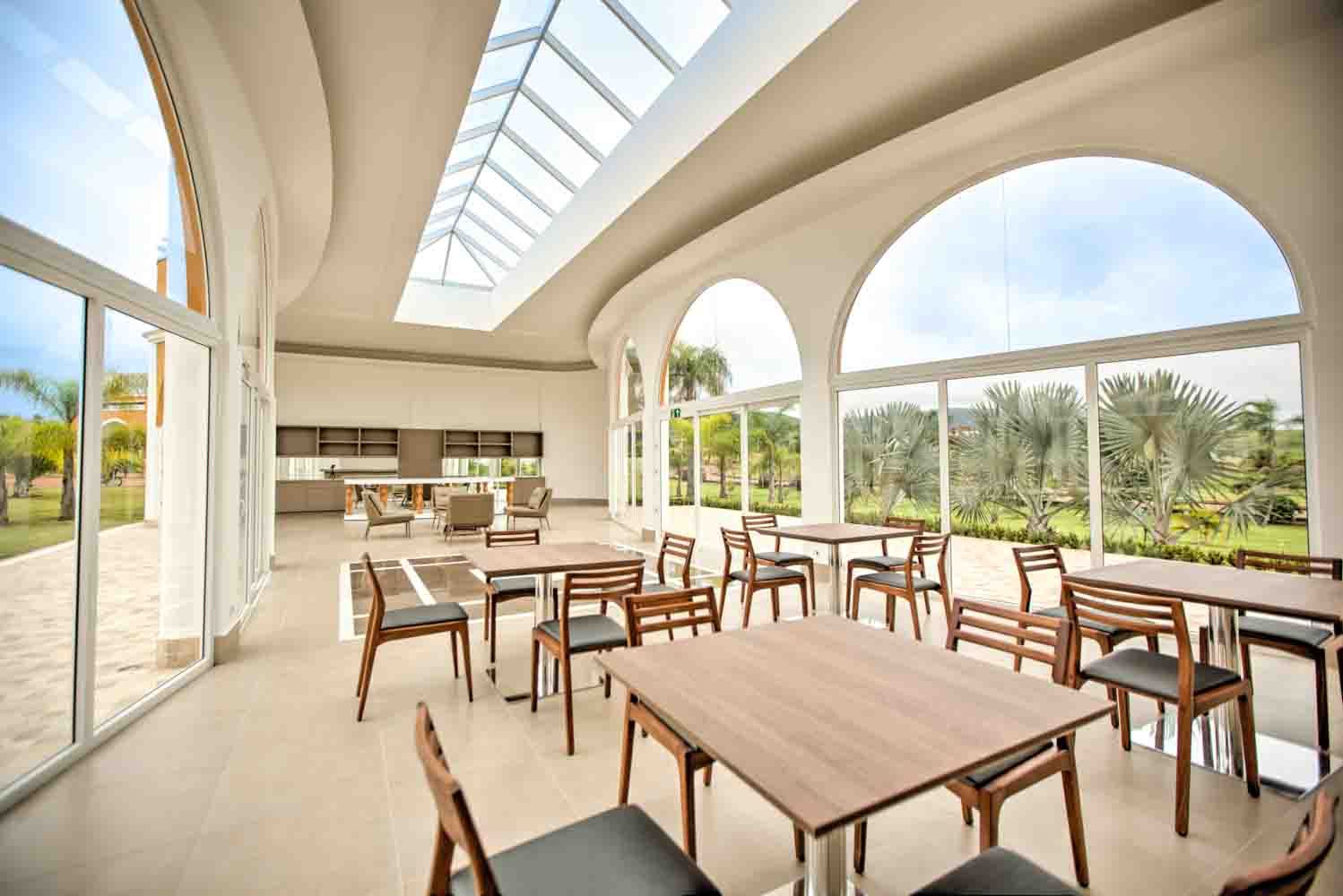 Interior de um espaço de jantar iluminado com mesas, cadeiras e grandes janelas em arco com vista para a área verde.