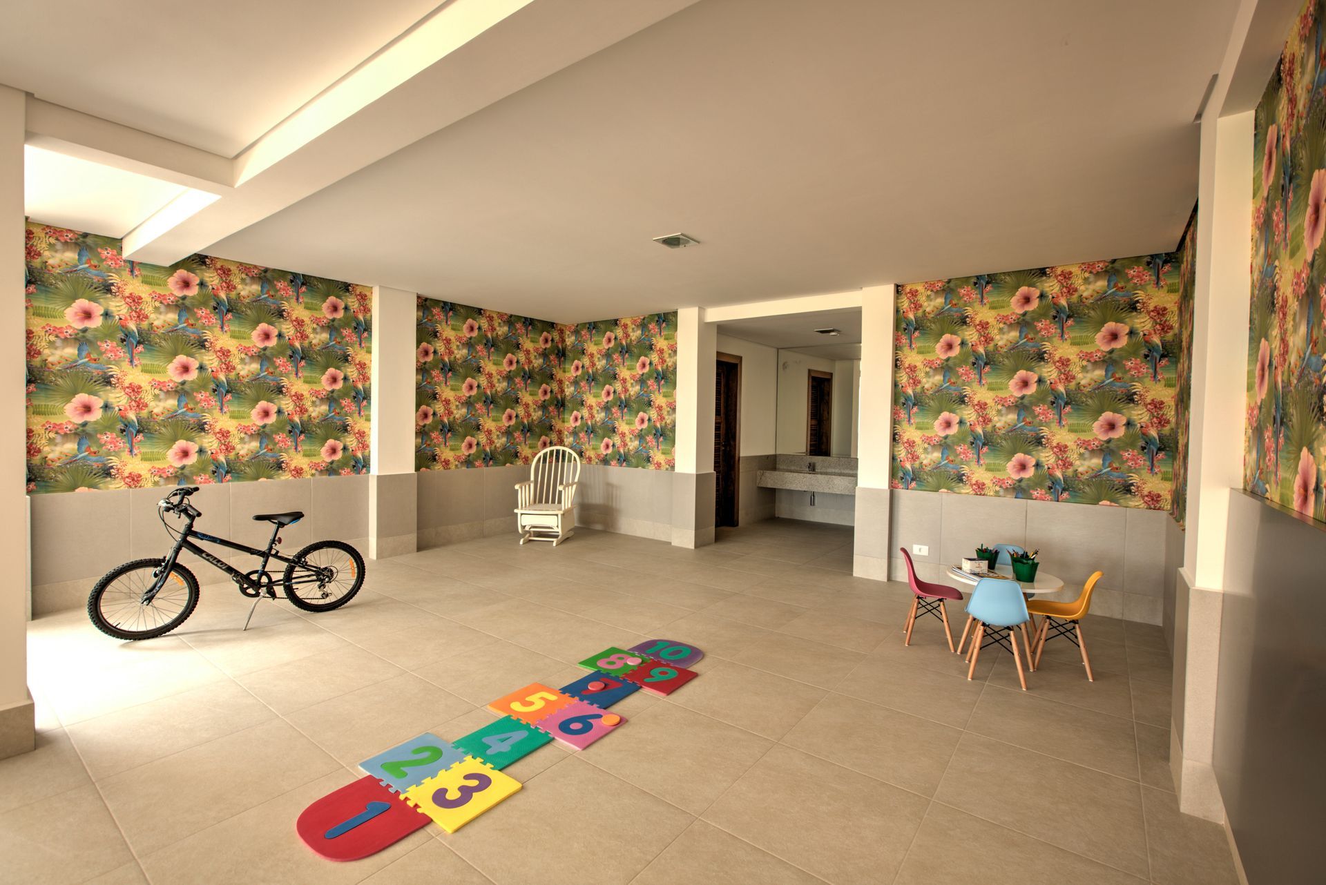 Sala de jogos com papel de parede floral, bicicleta, amarelinha e uma pequena mesa com cadeiras.