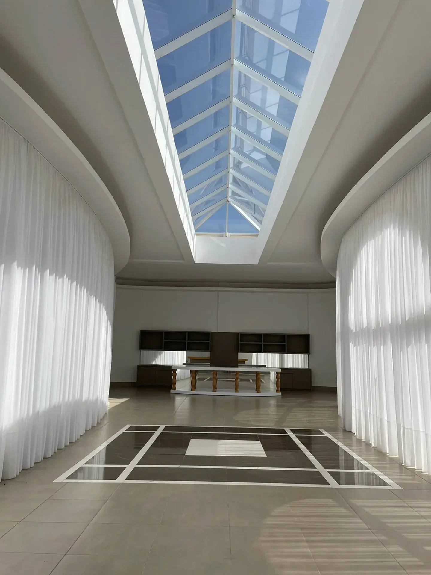 Sala comprida, com paredes brancas e teto de vidro. Cortinas decoram as laterais e os móveis ficam ao longe.