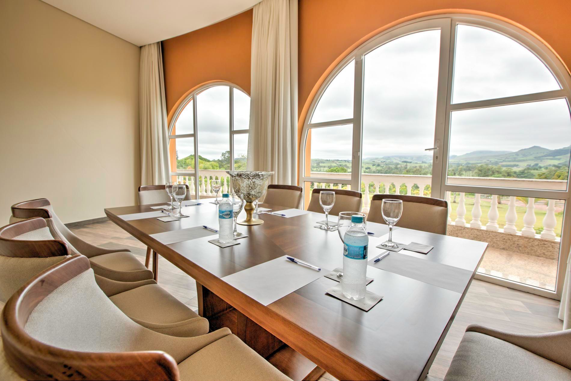Sala de conferências com grandes janelas em arco com vista para paisagem. Mesa e cadeiras marrons dispostas para uma reunião.