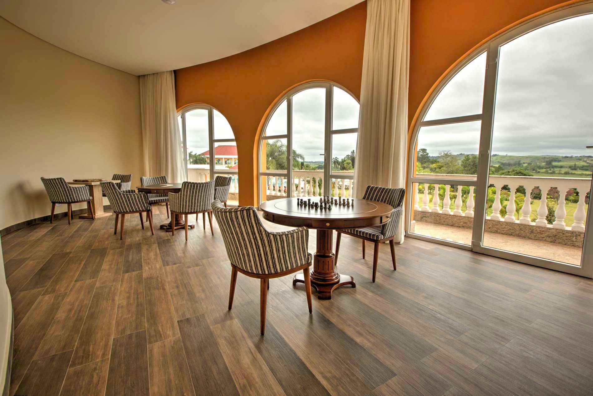 Sala com janelas em arco, mesas, cadeiras estampadas e varanda com vista para a paisagem. Piso de madeira, paredes laranja.