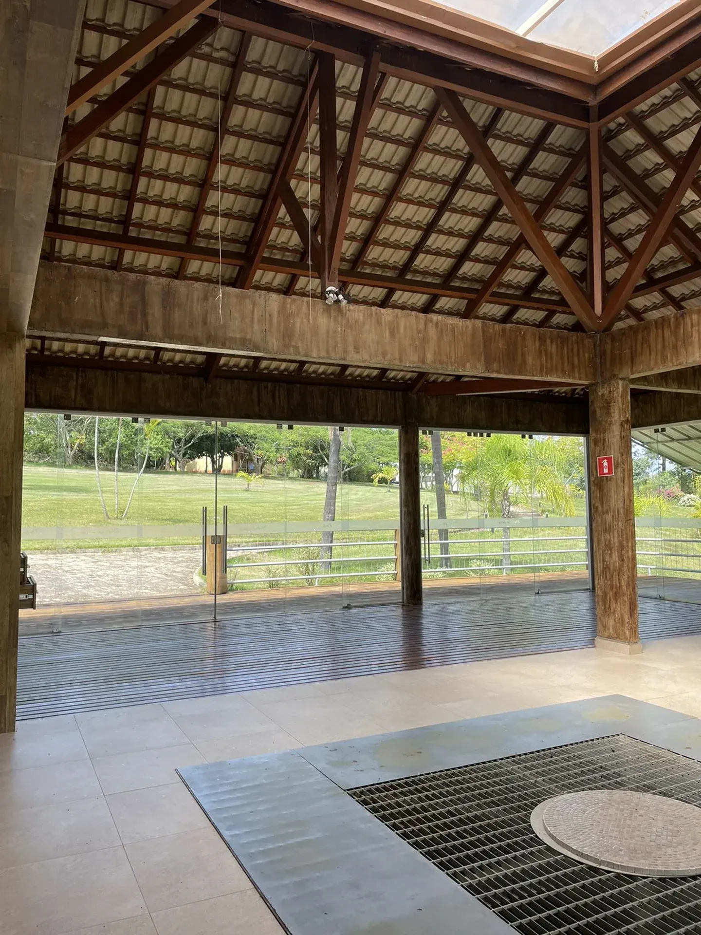 Pátio externo coberto com piso de cerâmica, vigas de madeira e vista para um campo verde.