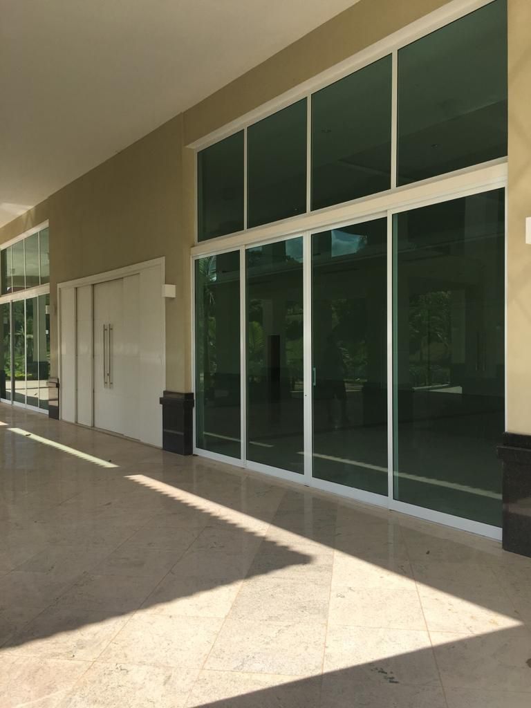 Corredor externo com janelas grandes, porta branca, paredes bege e luz solar.