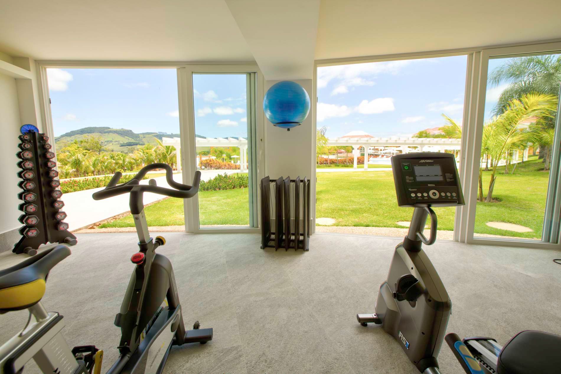 Academia com bicicletas ergométricas e pesos, com vista para um gramado verde e para o mar.