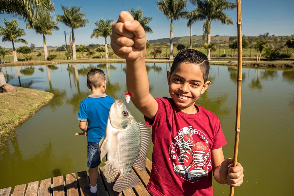 Menino segura orgulhosamente o peixe que pescou, sorrindo perto de um lago; outro menino pesca atrás dele.