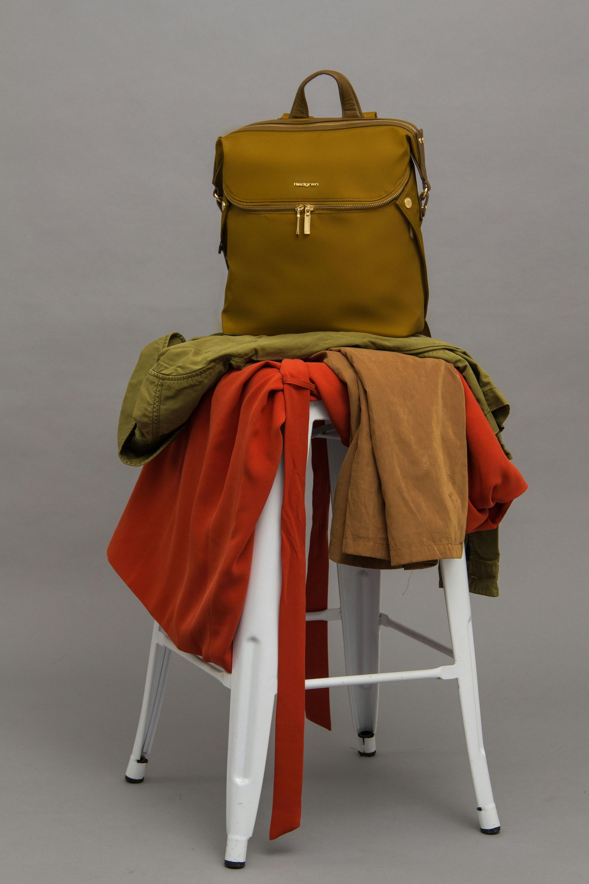 Uma mochila verde em cima de um banquinho branco com roupas laranja e marrons sobre um fundo cinza.