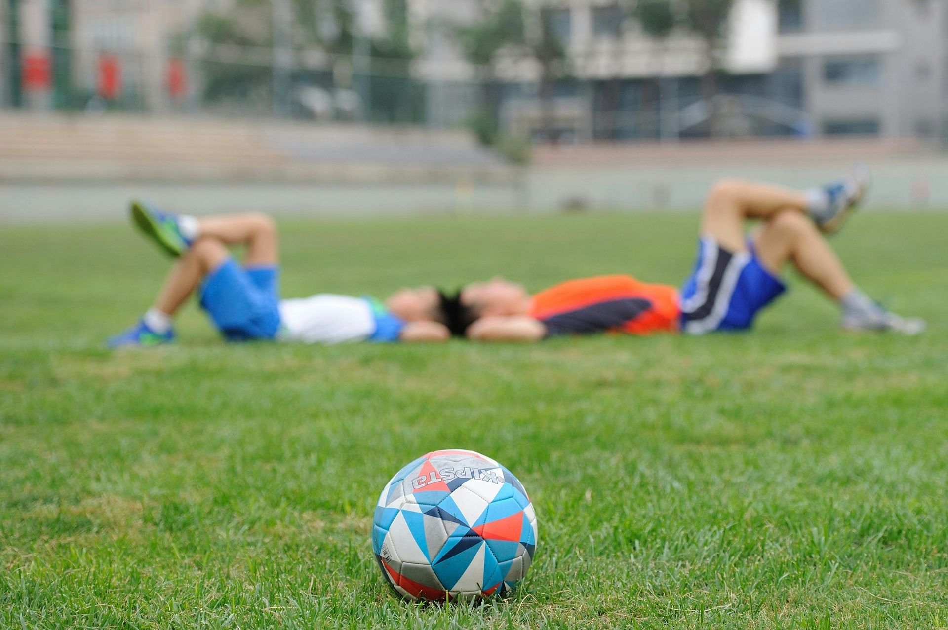 Bola de futebol na grama verde com duas pessoas descansando ao fundo.
