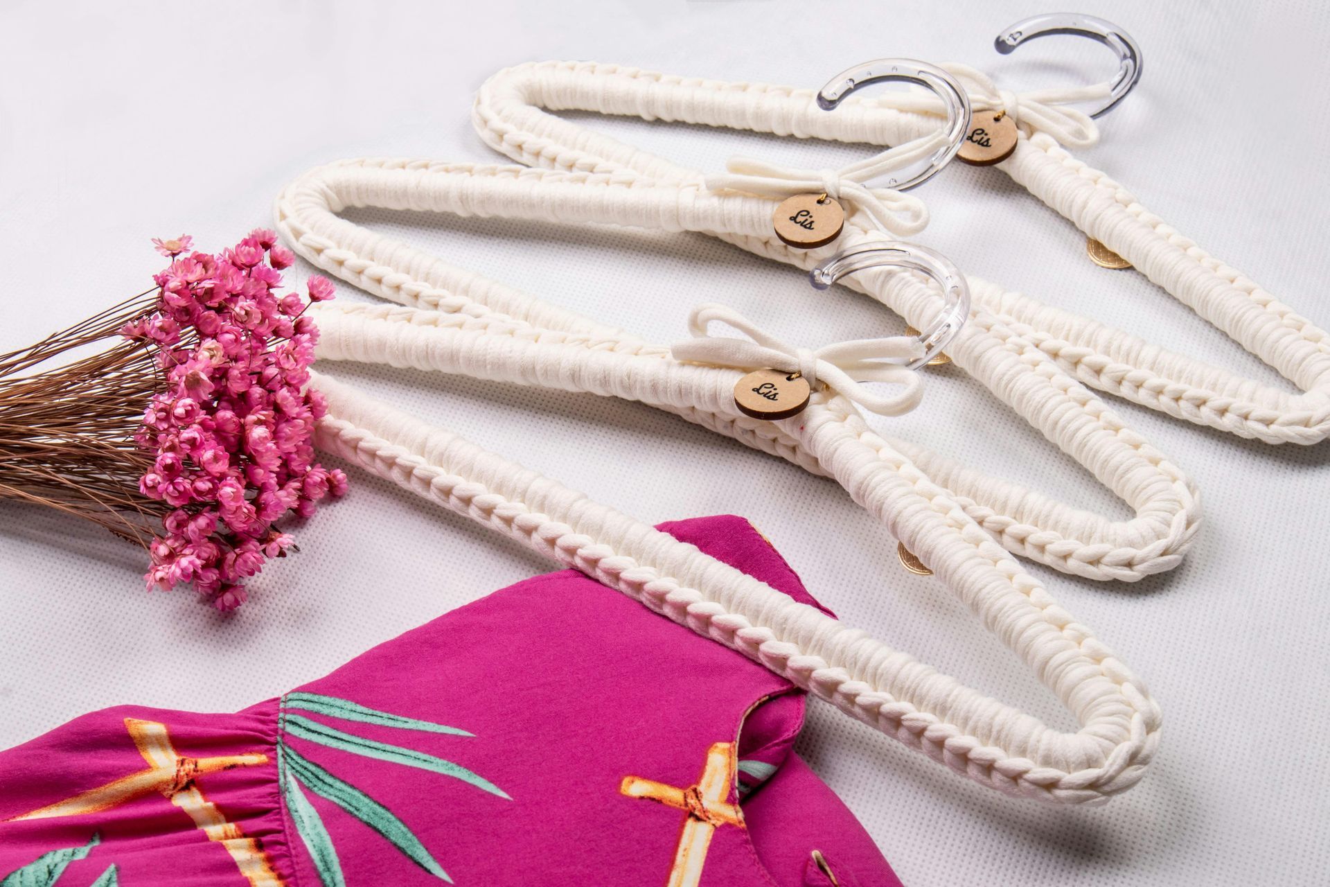 Três cabides brancos de crochê com botões de madeira e um vestido rosa, flores sobre superfície branca.