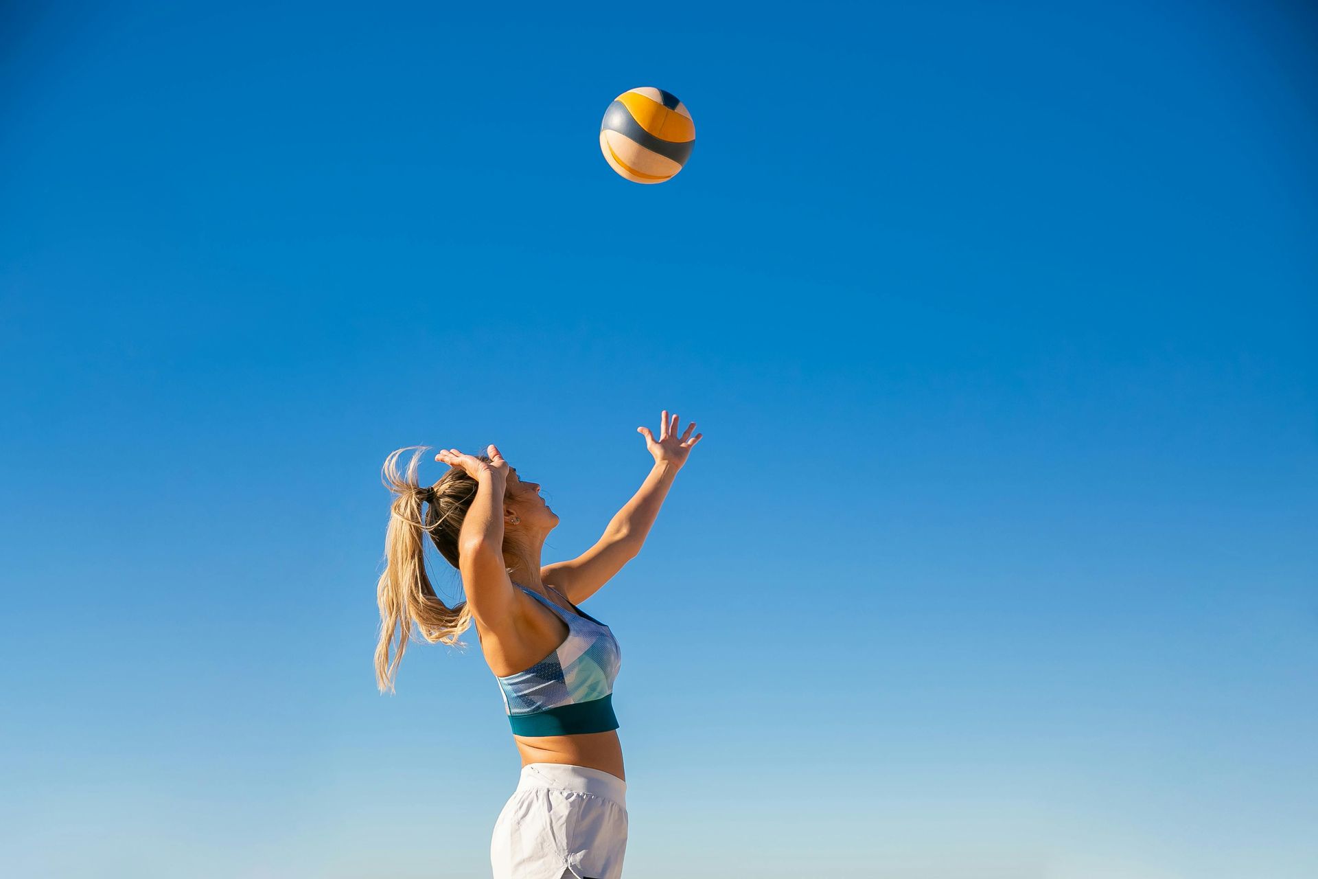 Mulher com roupa esportiva se levantando para rebater uma bola de vôlei contra um céu azul.