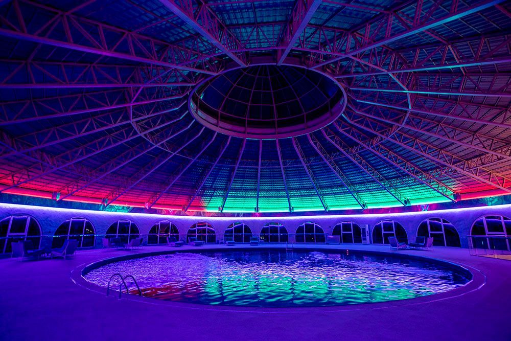 Piscina coberta sob uma cúpula colorida, com iluminação roxa, verde e rosa refletindo na água.