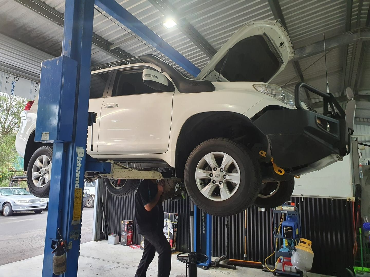 White SUV raised on a lift in a repair shop. Mechanic working underneath. Hood open. — Adam's Automotive Centre in Nambour, QLD