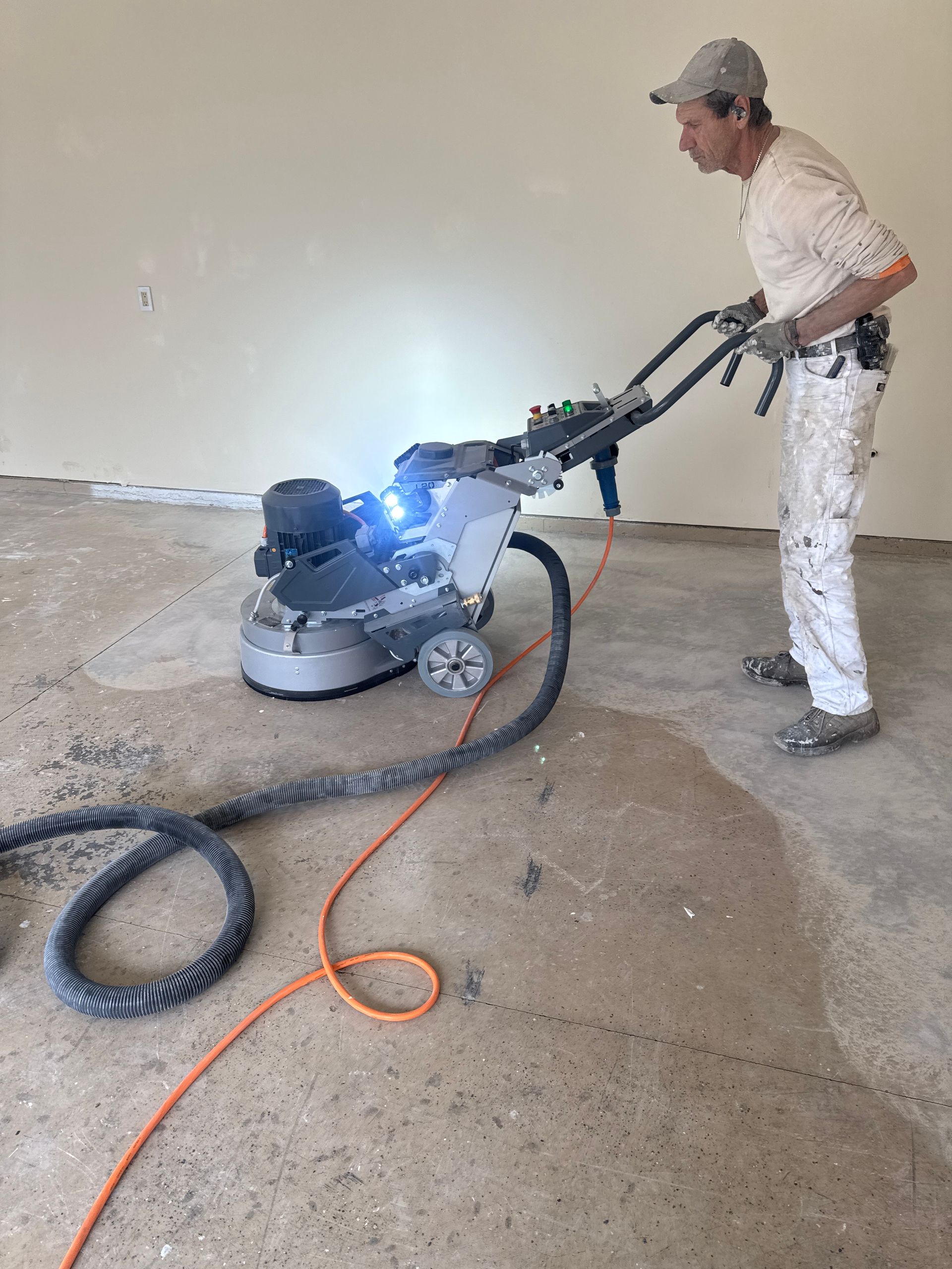 Man using a concrete grinder on a floor, wearing white work clothes. Indoors, grey and orange cords.