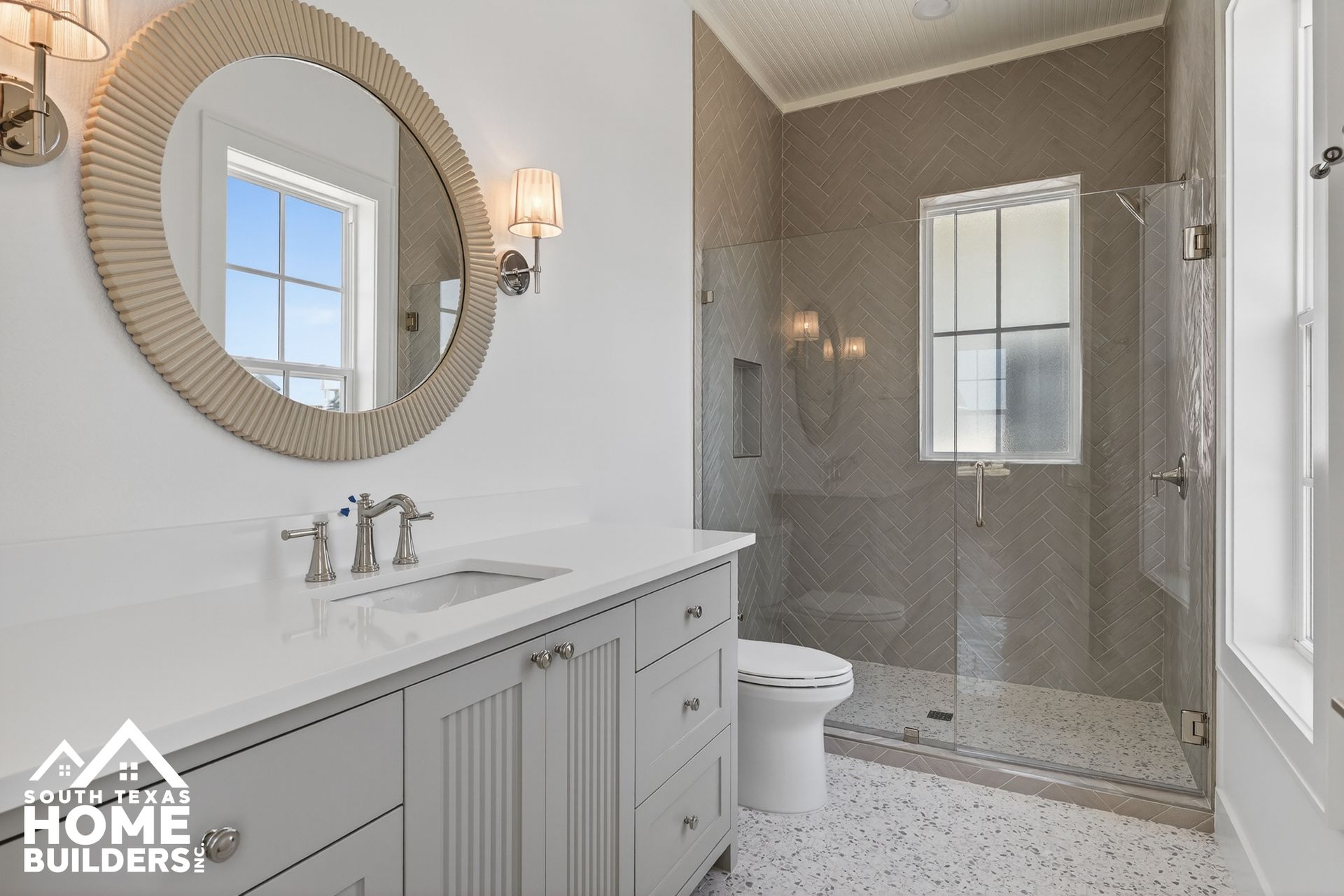 Bright bathroom with white vanity, round mirror, and glass-enclosed shower with gray tile.