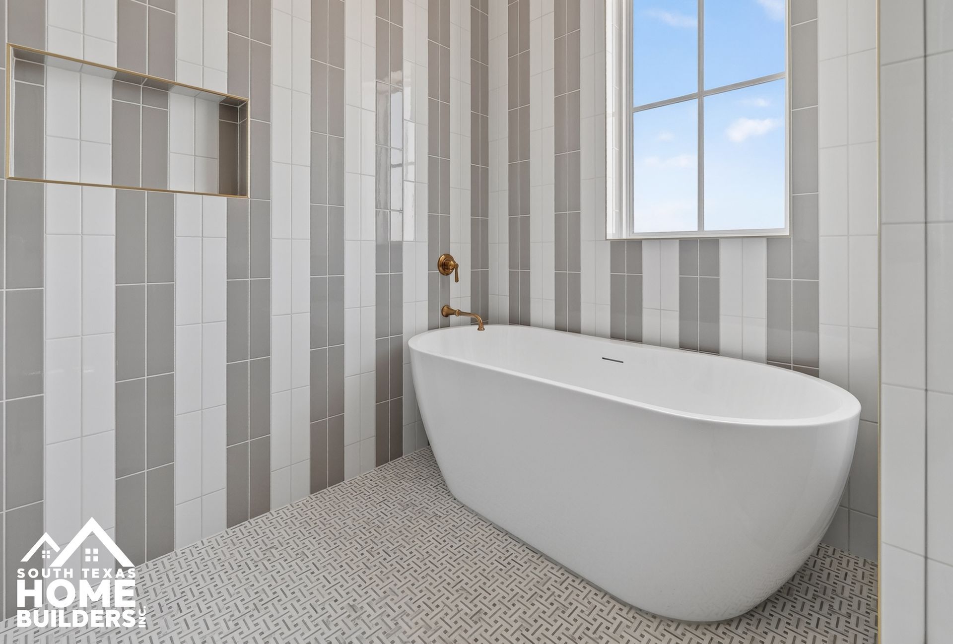 Modern white bathroom with freestanding tub, striped tile walls, pebble floor, and a window