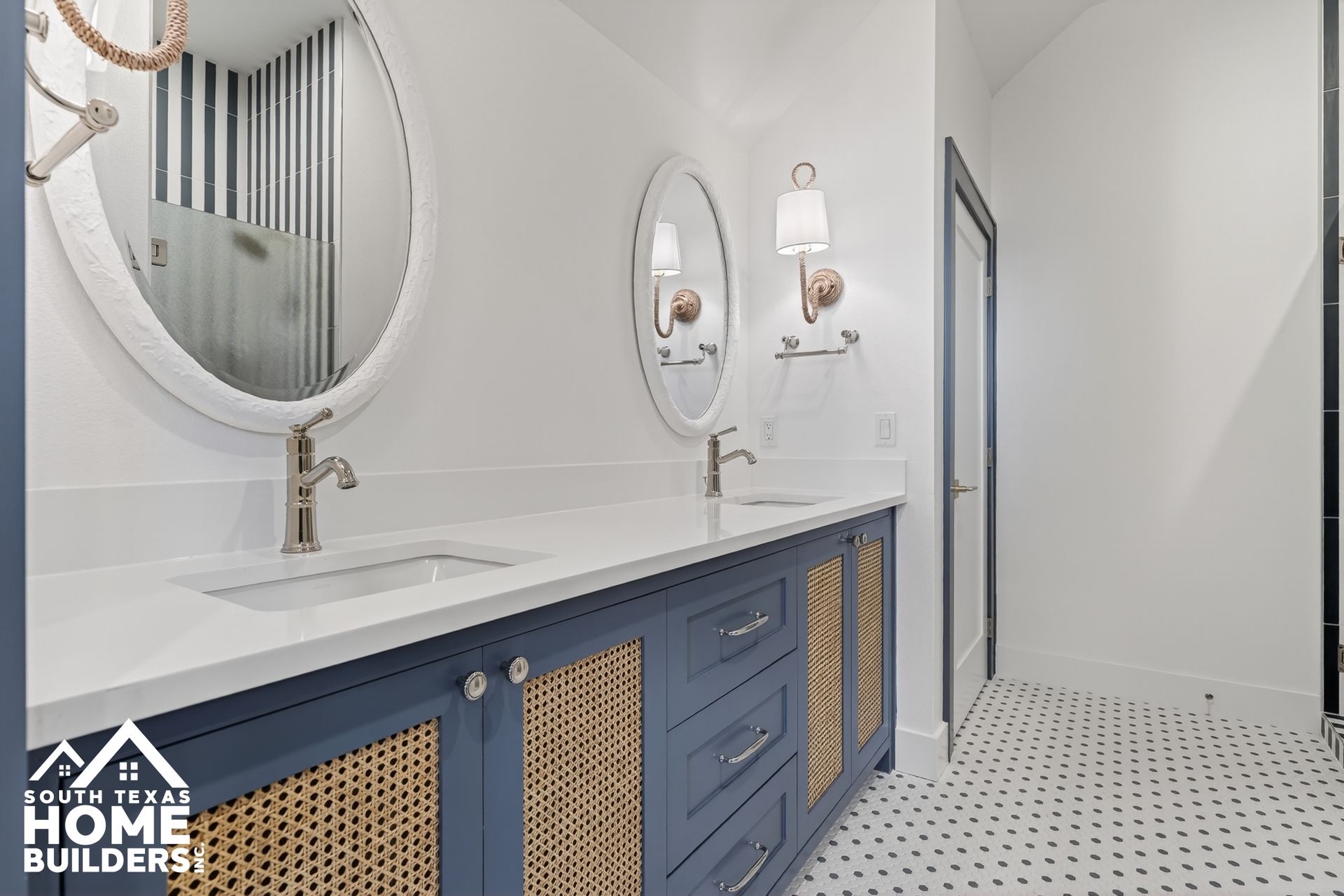 Modern bathroom with blue vanity, oval mirrors, white tile floor, and woven cabinet panels