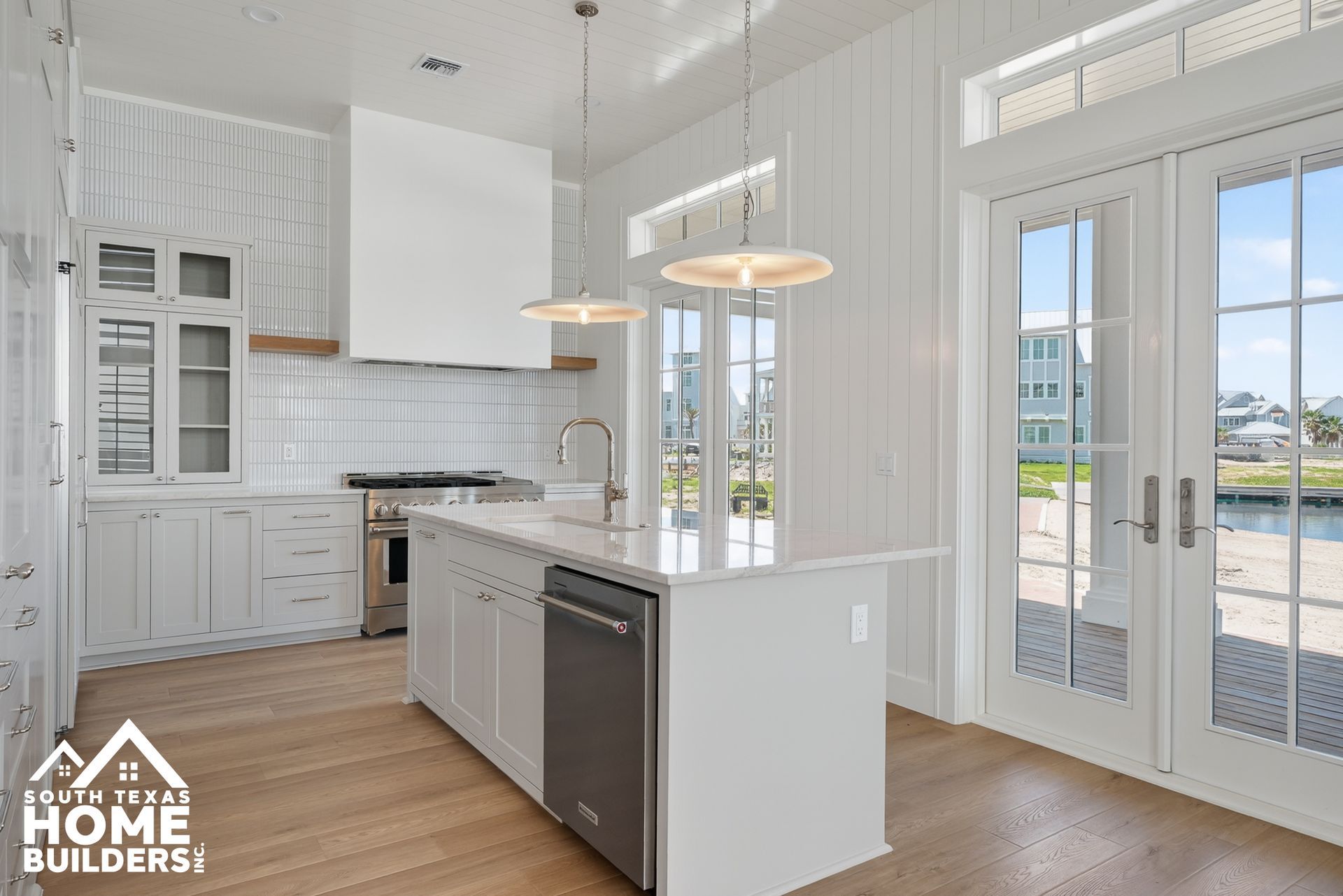 Bright white kitchen with island, pendant lights, wood floors, and French doors to outside.