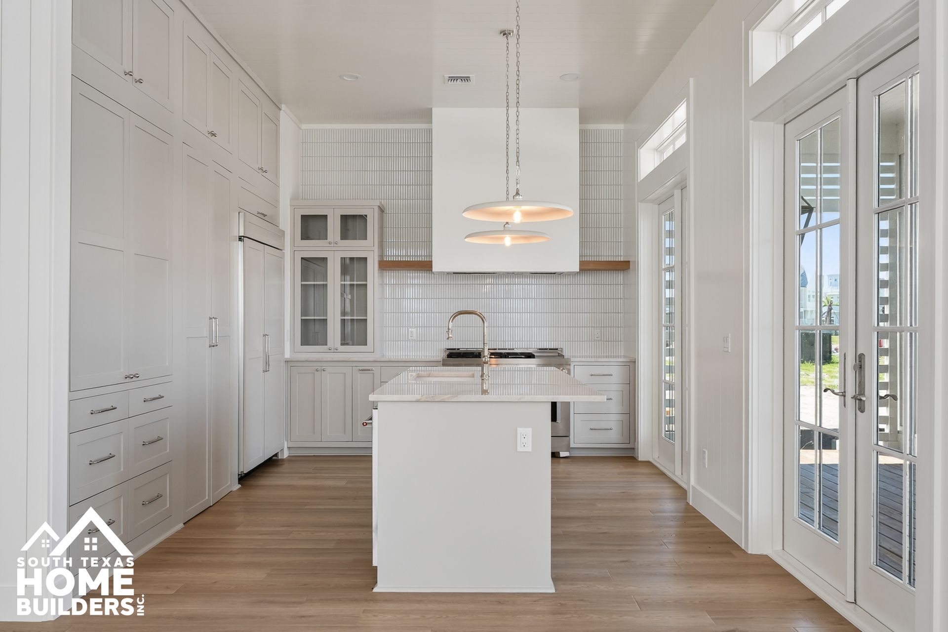 Bright white modern kitchen with island, wood floors, tall cabinets, and large windows