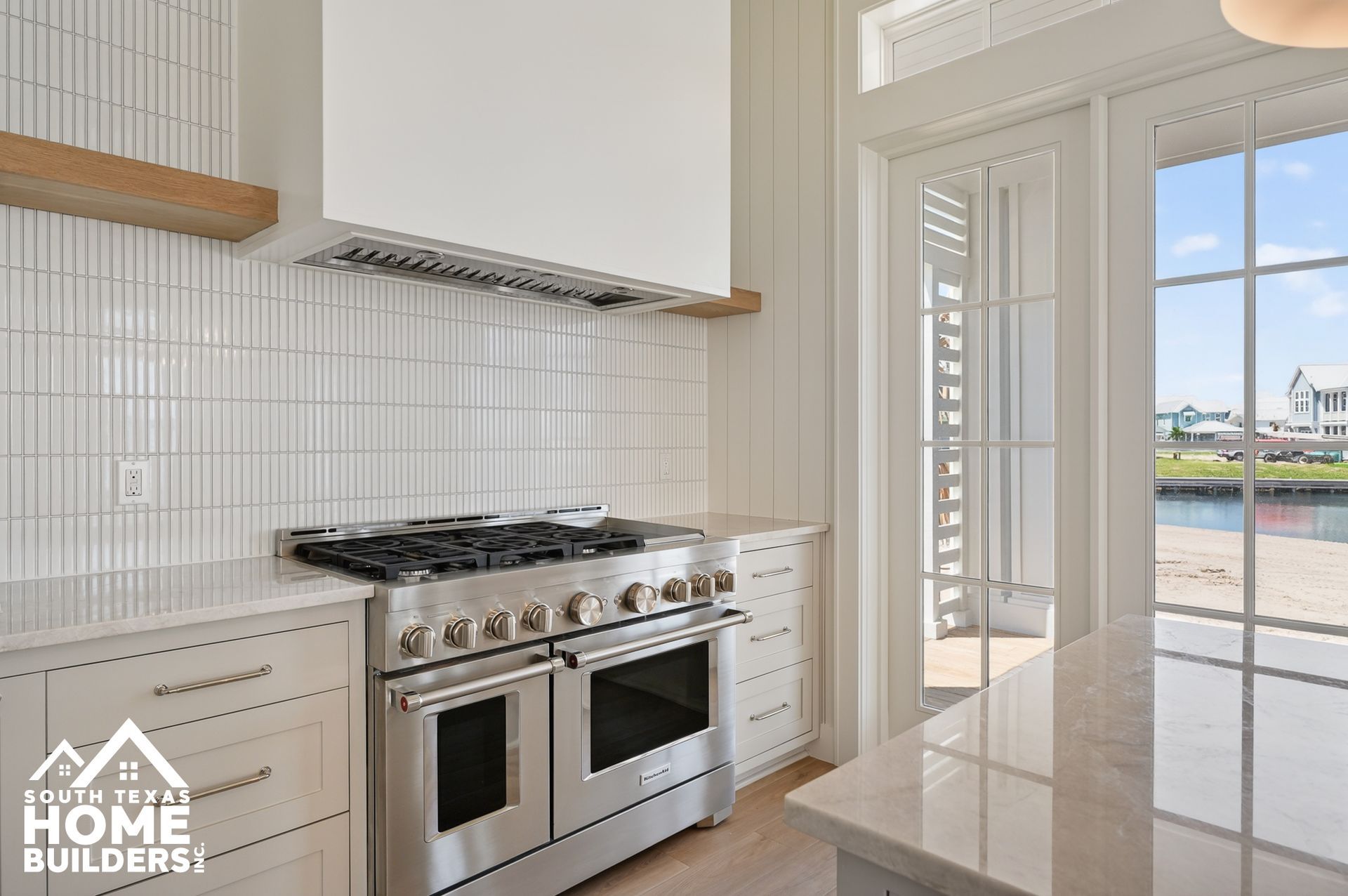 Modern white kitchen with stainless steel stove, glossy tile, and glass doors opening to a water view