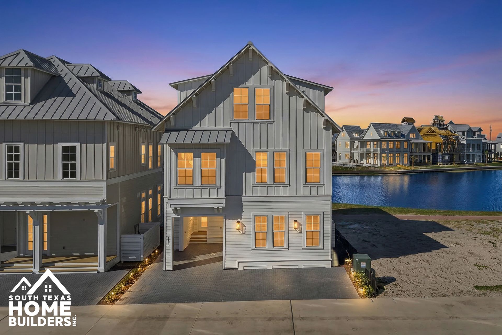 Modern gray waterfront homes at dusk, with warm lit windows and a calm harbor in the background