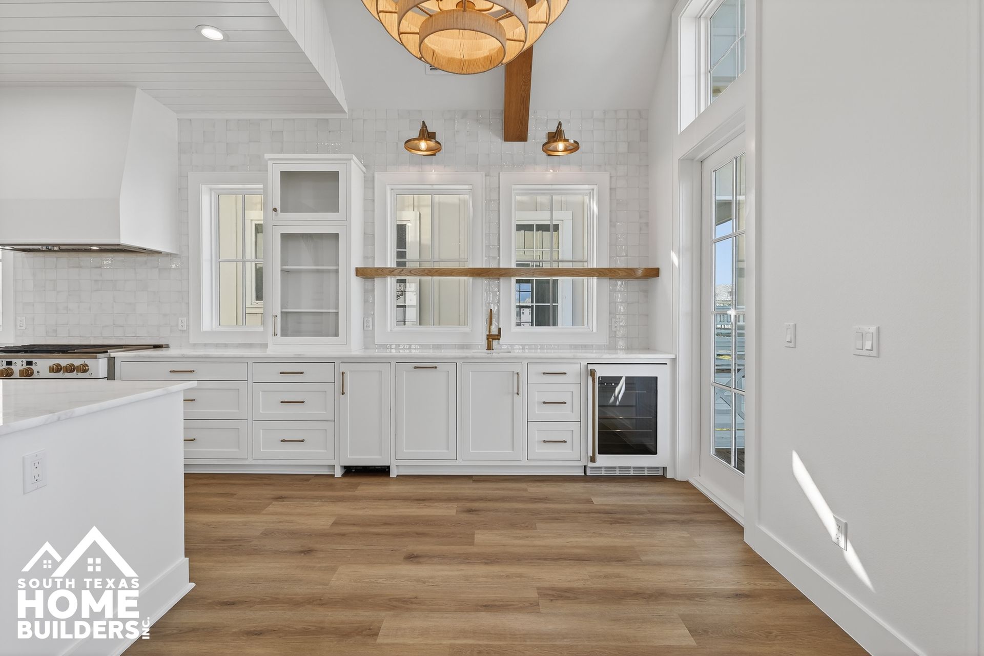 Bright white modern kitchen with wood floors, island, brass pendants, and stainless appliances