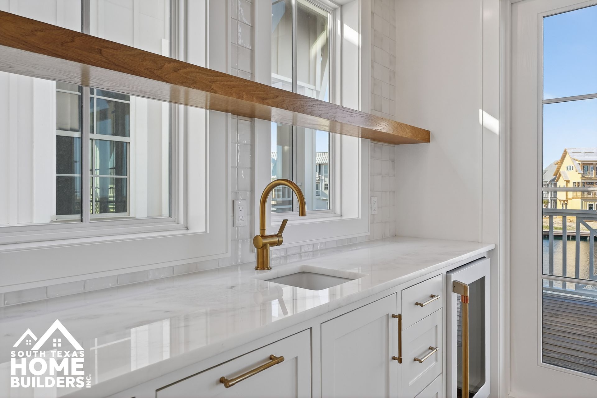 Bright modern kitchen sink with gold faucet, white cabinets, and large windows overlooking a balcony.