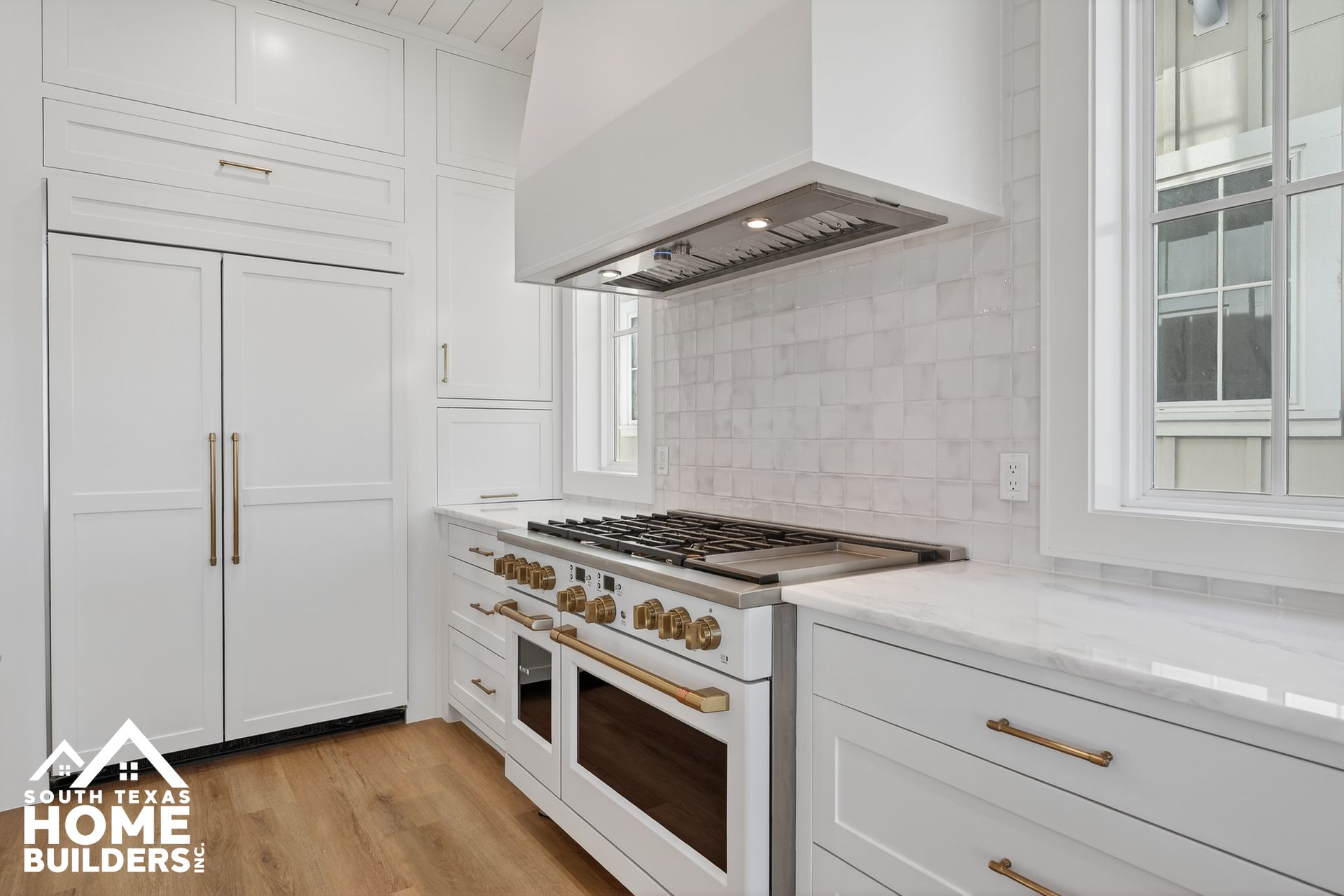 Bright white kitchen with gold hardware, stainless range hood, marble backsplash, and wood floors