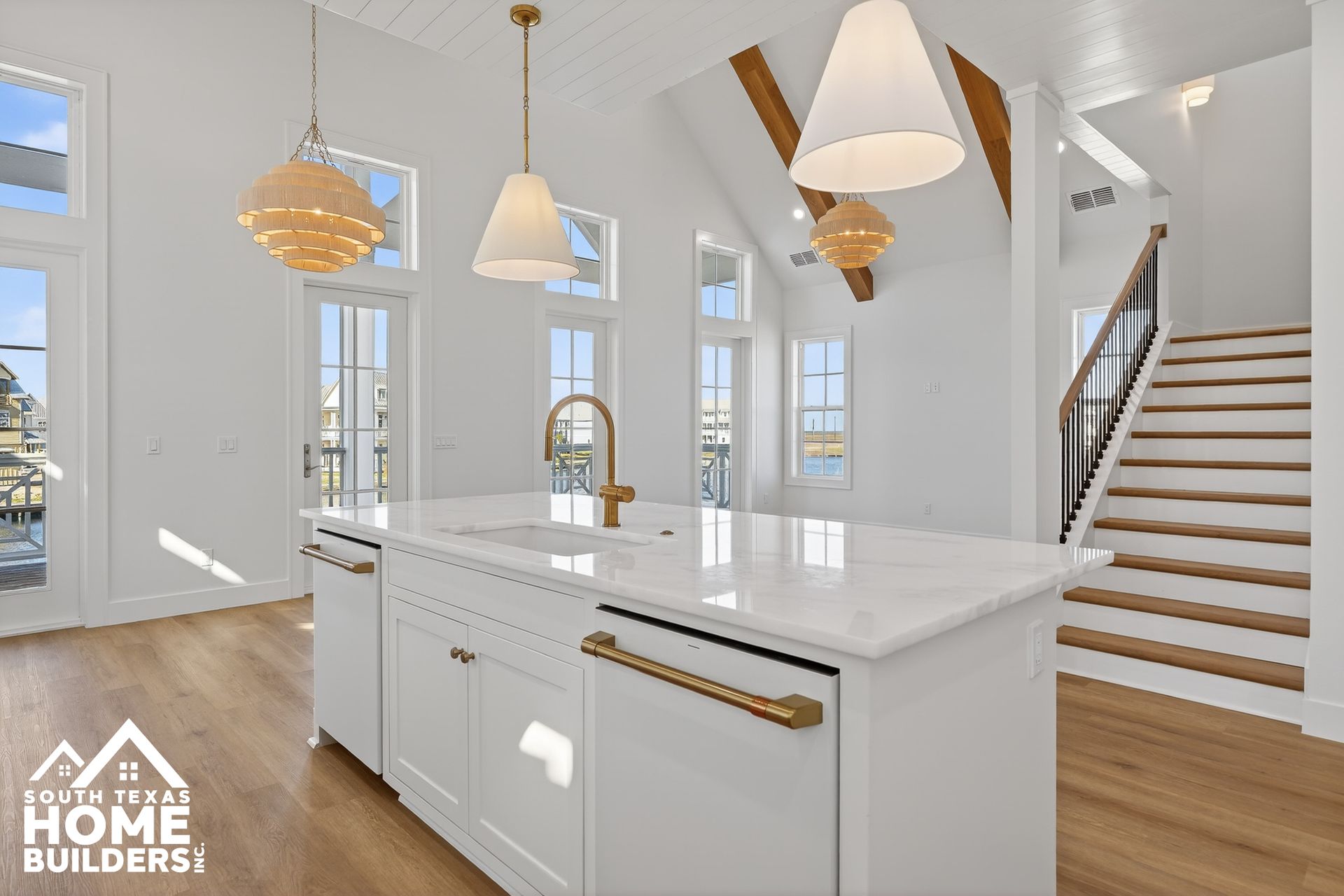 Bright white modern kitchen with island, gold fixtures, wood floors, and staircase in the background
