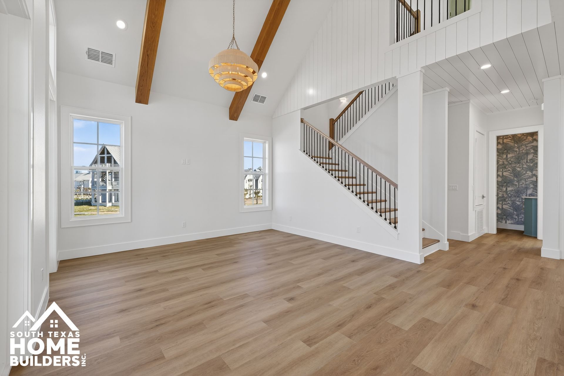 Bright open living room with vaulted white ceiling, wood beams, staircase, and light wood floors