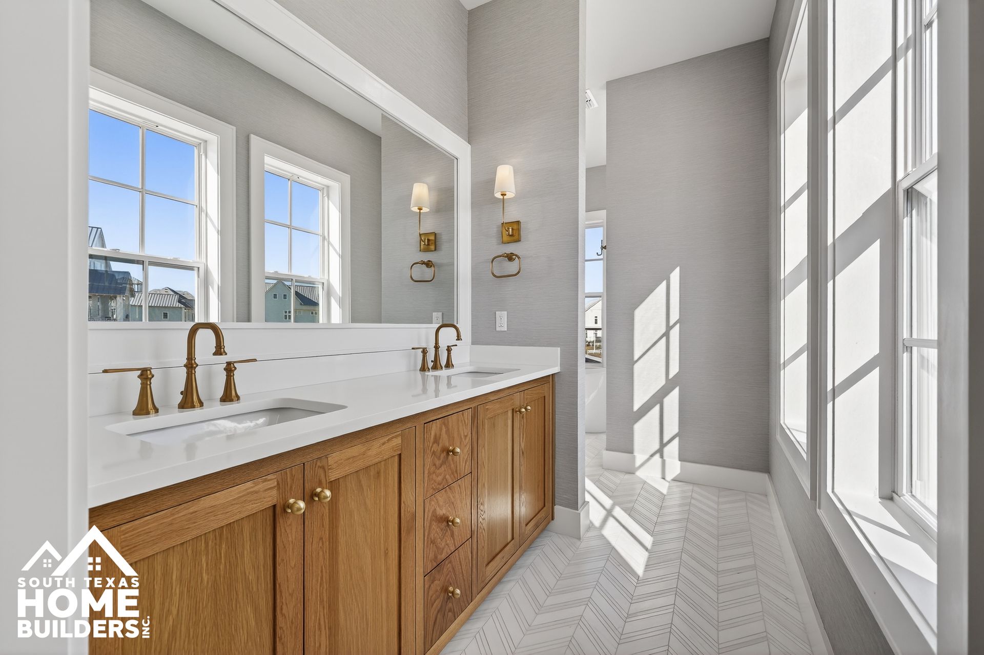 Bright bathroom with white marble vanity, wood cabinets, brass faucets, and large sunlit windows