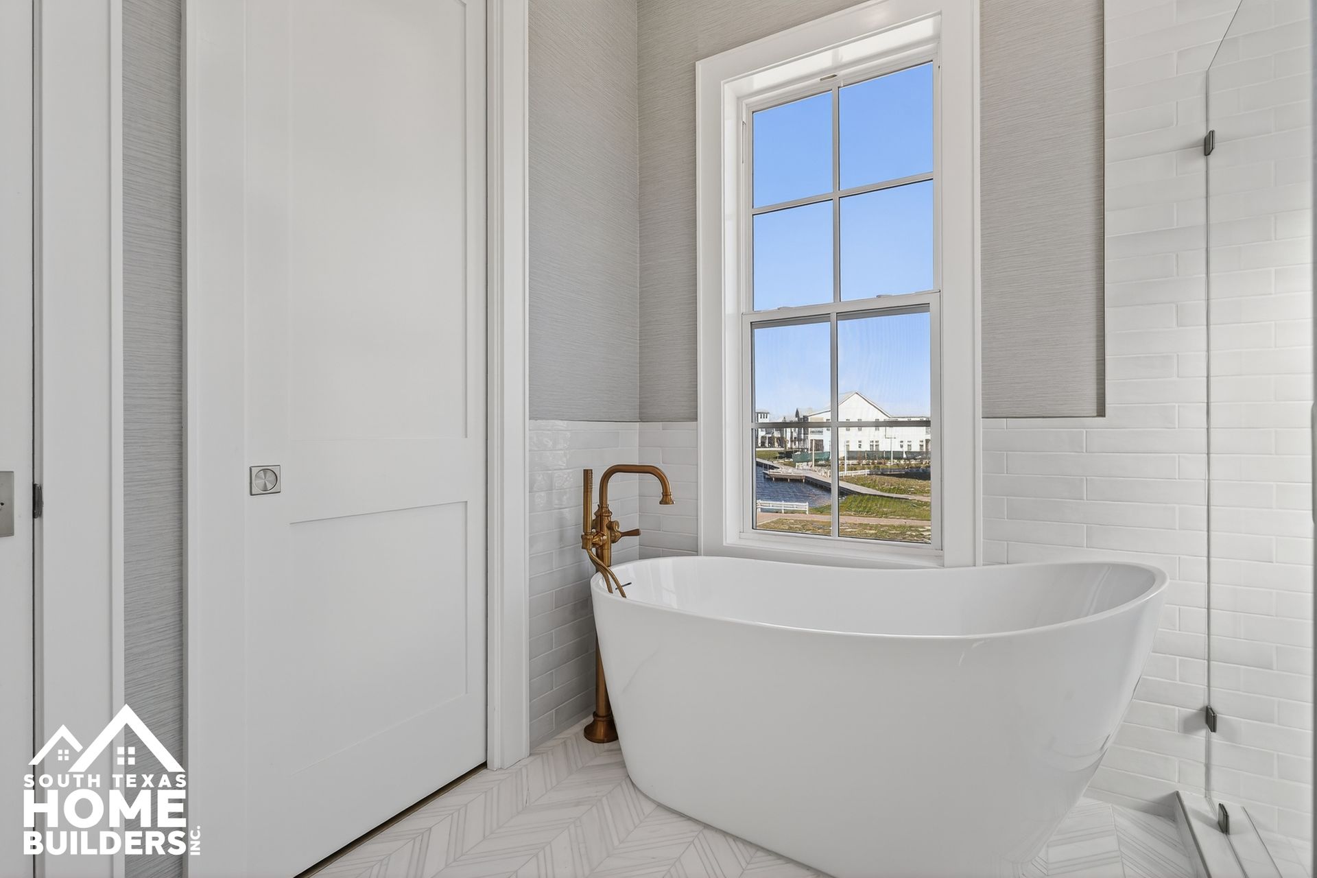 Modern white bathroom with freestanding tub, gold faucet, and window view of a waterfront home
