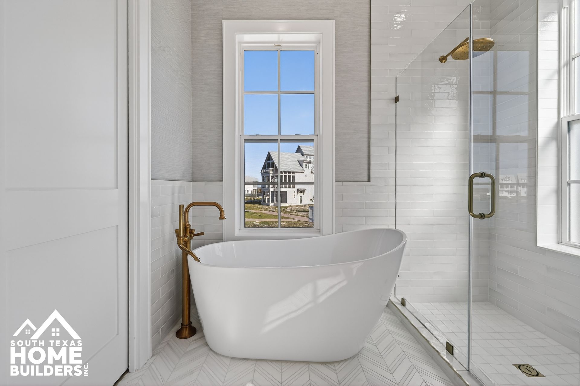 Bright modern bathroom with a freestanding tub, glass shower, gold fixtures, and a window view.