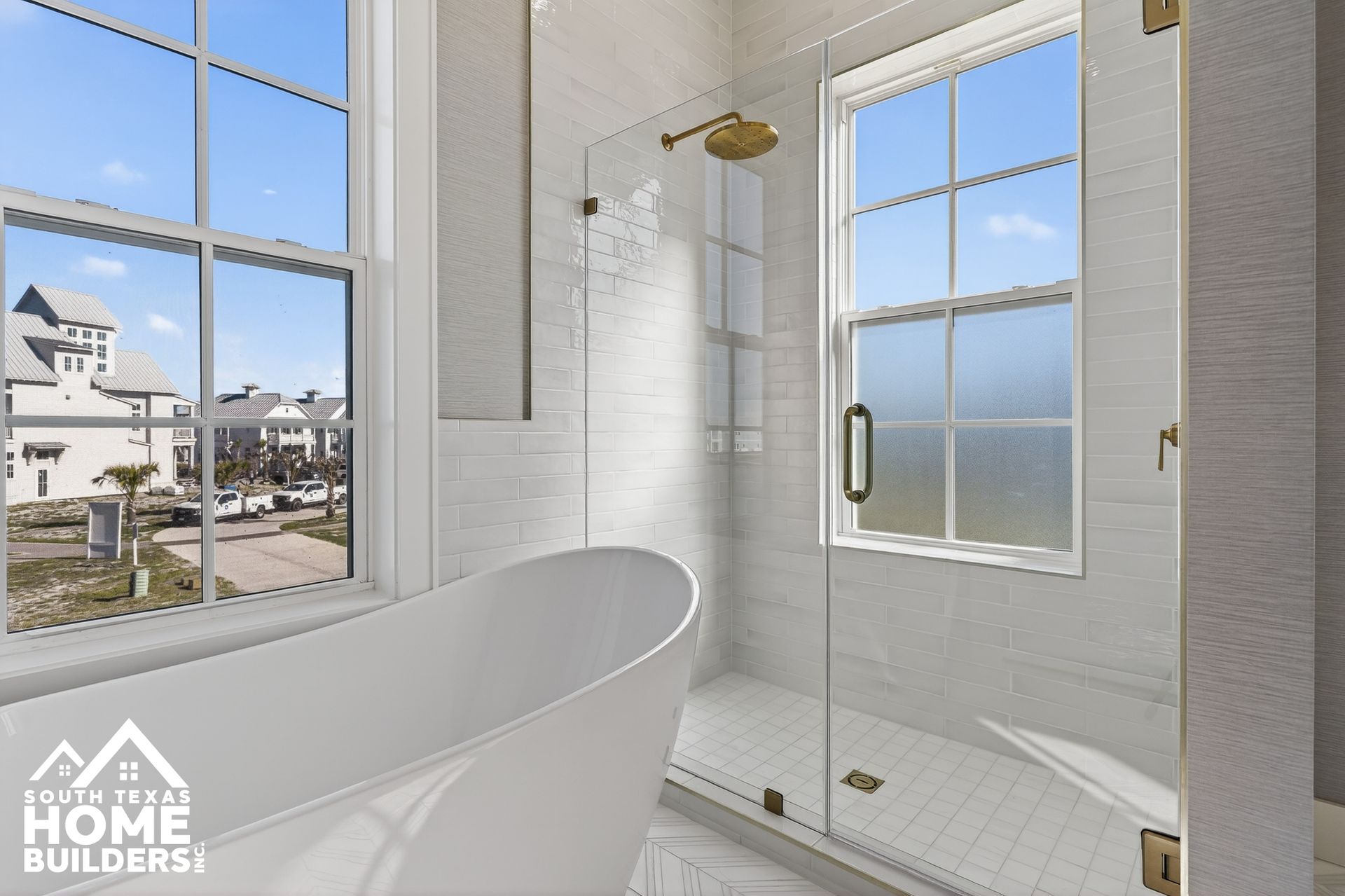 Bright bathroom with freestanding tub, glass shower, and large windows overlooking houses and water