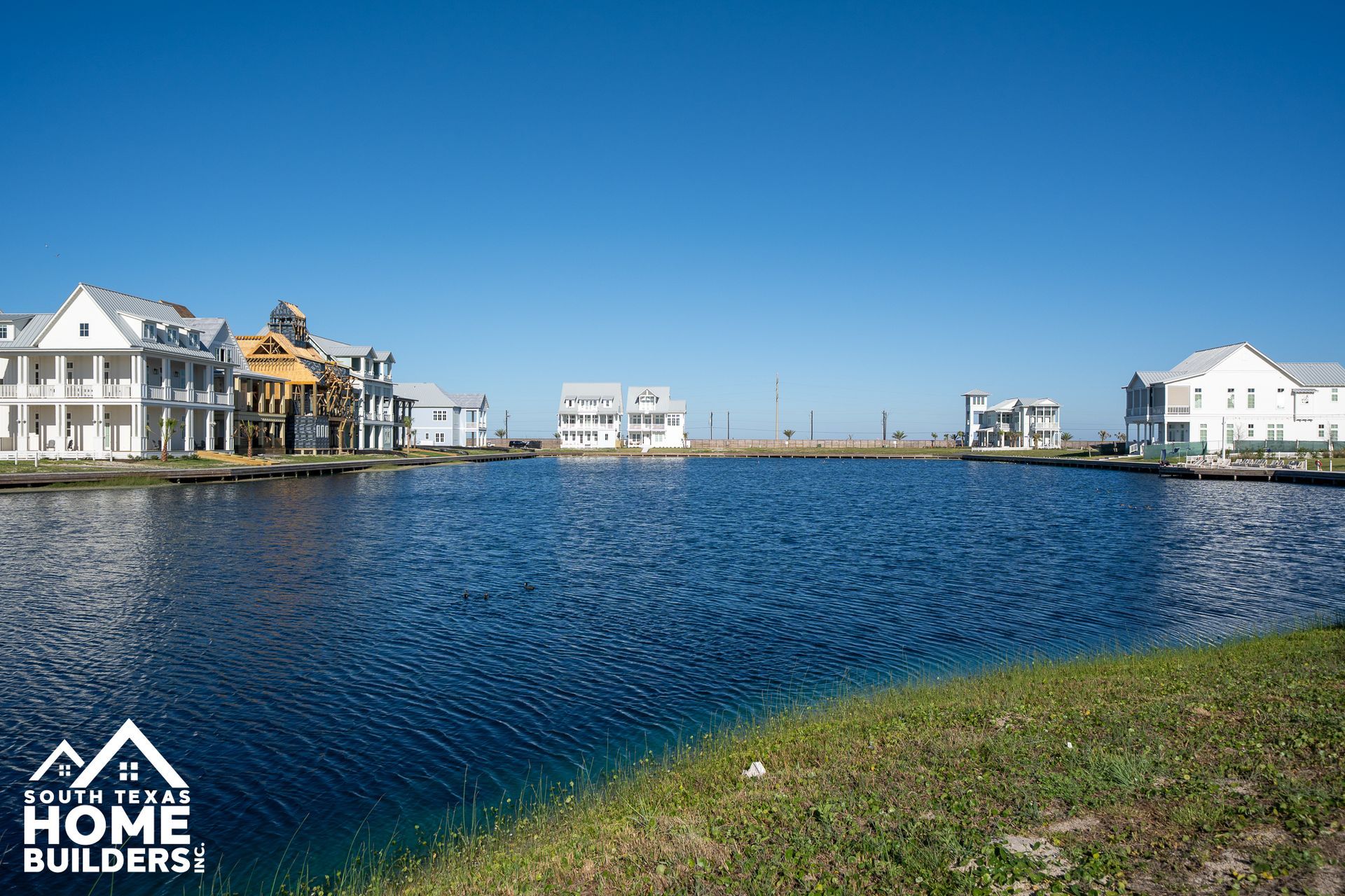 Waterfront homes beside a calm blue lagoon under a clear sky, with a HOME BUILDERS logo in the corner